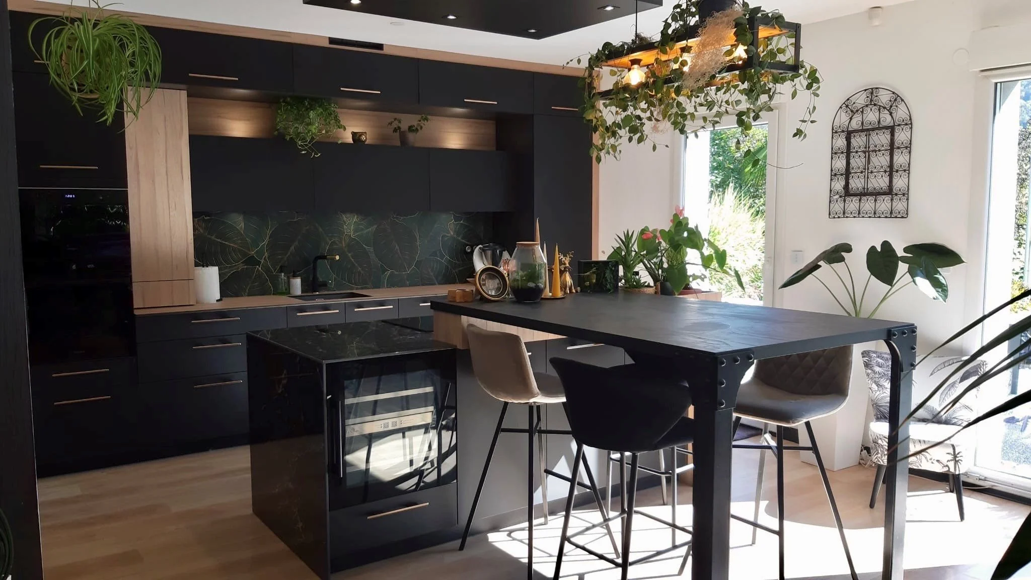 Cuisine moderne avec meubles noirs, plan de travail en bois, il y a des plantes et une table avec des chaises autour, lumière naturelle par les fenêtres.