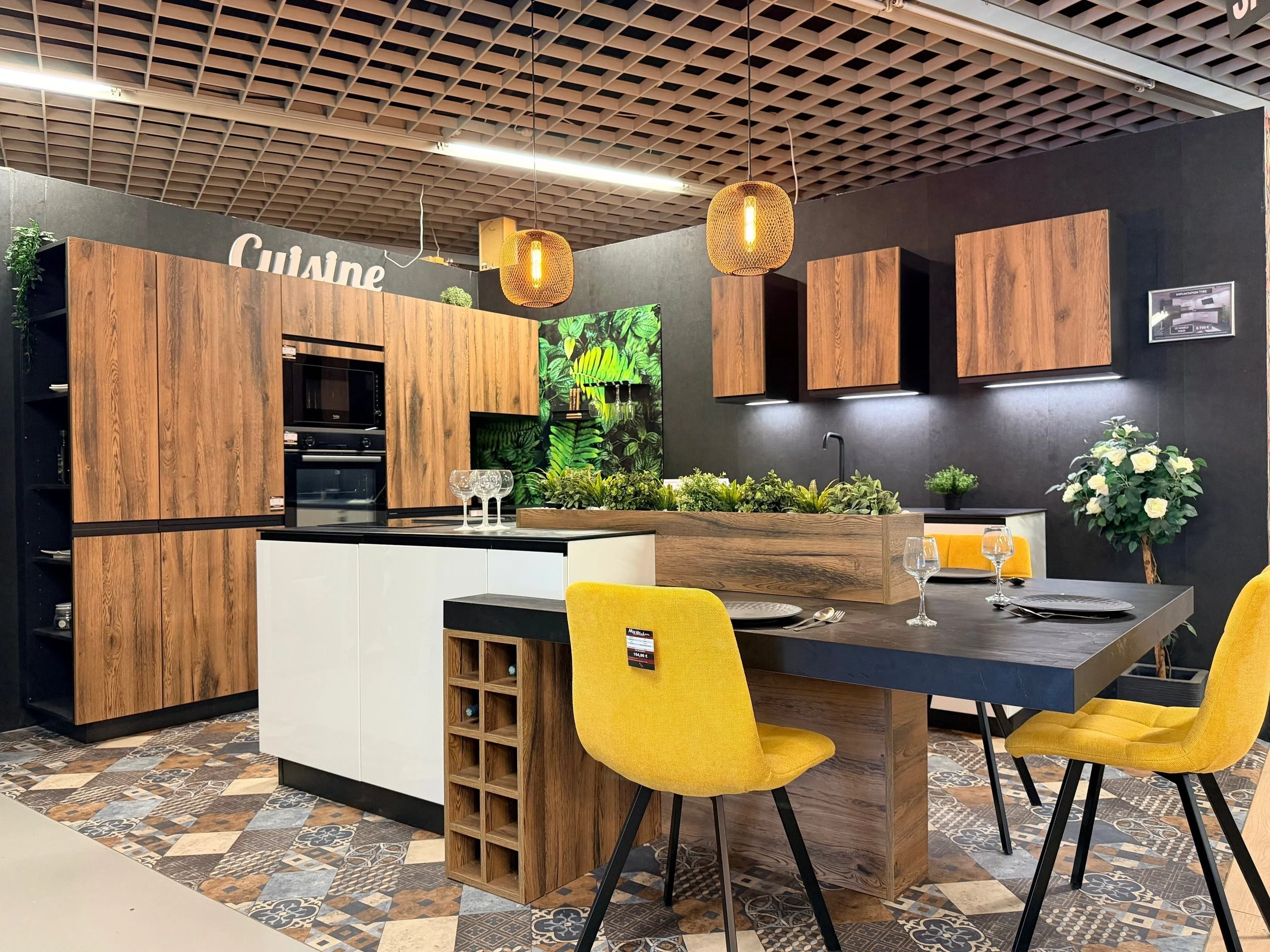 Une cuisine moderne avec des armoires en bois, un plan de travail noir, un îlot central blanc, une table à manger avec des chaises jaunes, et des décorations vertes, éclairée par deux lampes suspendues