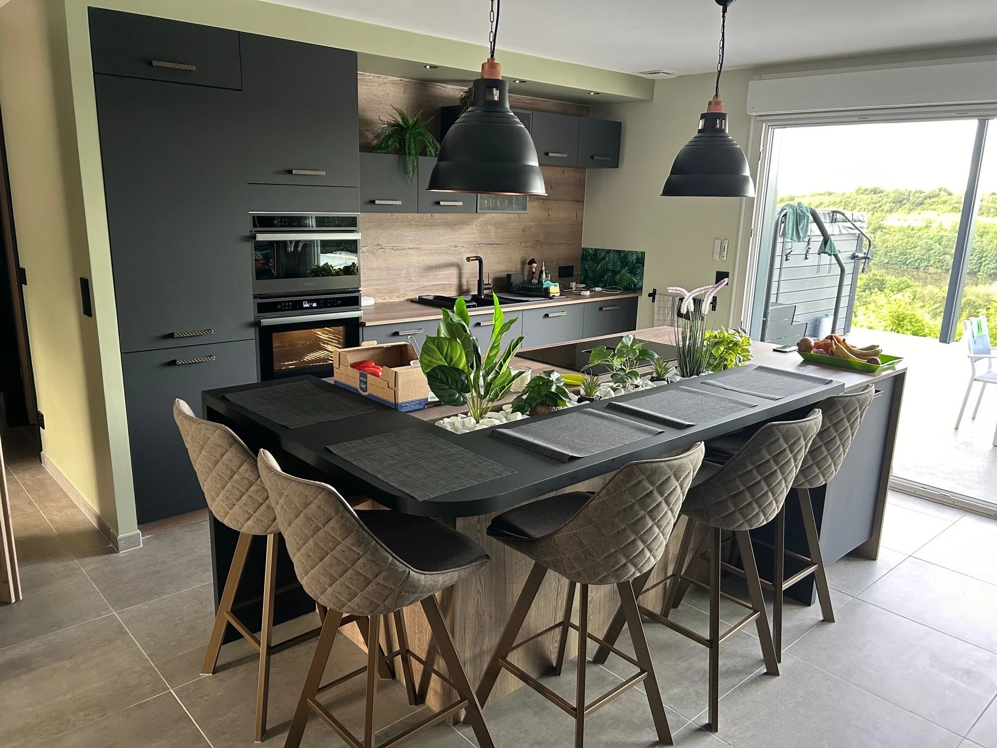 Cuisine moderne avec plan de travail central, chaises rembourrées, armoires grises, éclairages suspendus noirs, plantes vertes, grande fenêtre offrant vue sur la nature.