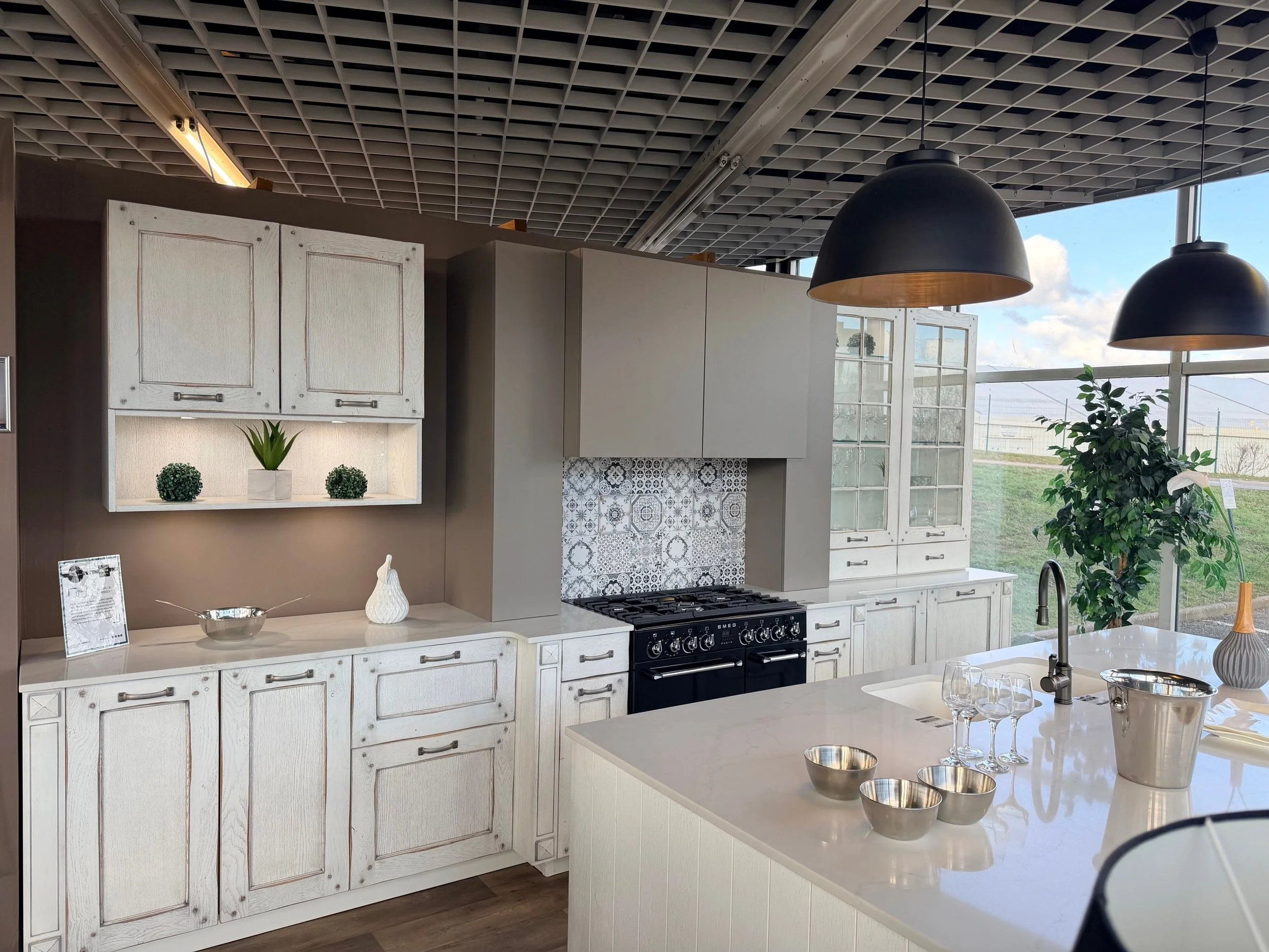 Cuisine moderne avec des armoires blanches, un plan de travail en marbre blanc, une cuisinière noire, deux lampes suspendues noires, des plantes et un grand mur de fenêtres laissant entrer la lumière naturelle.