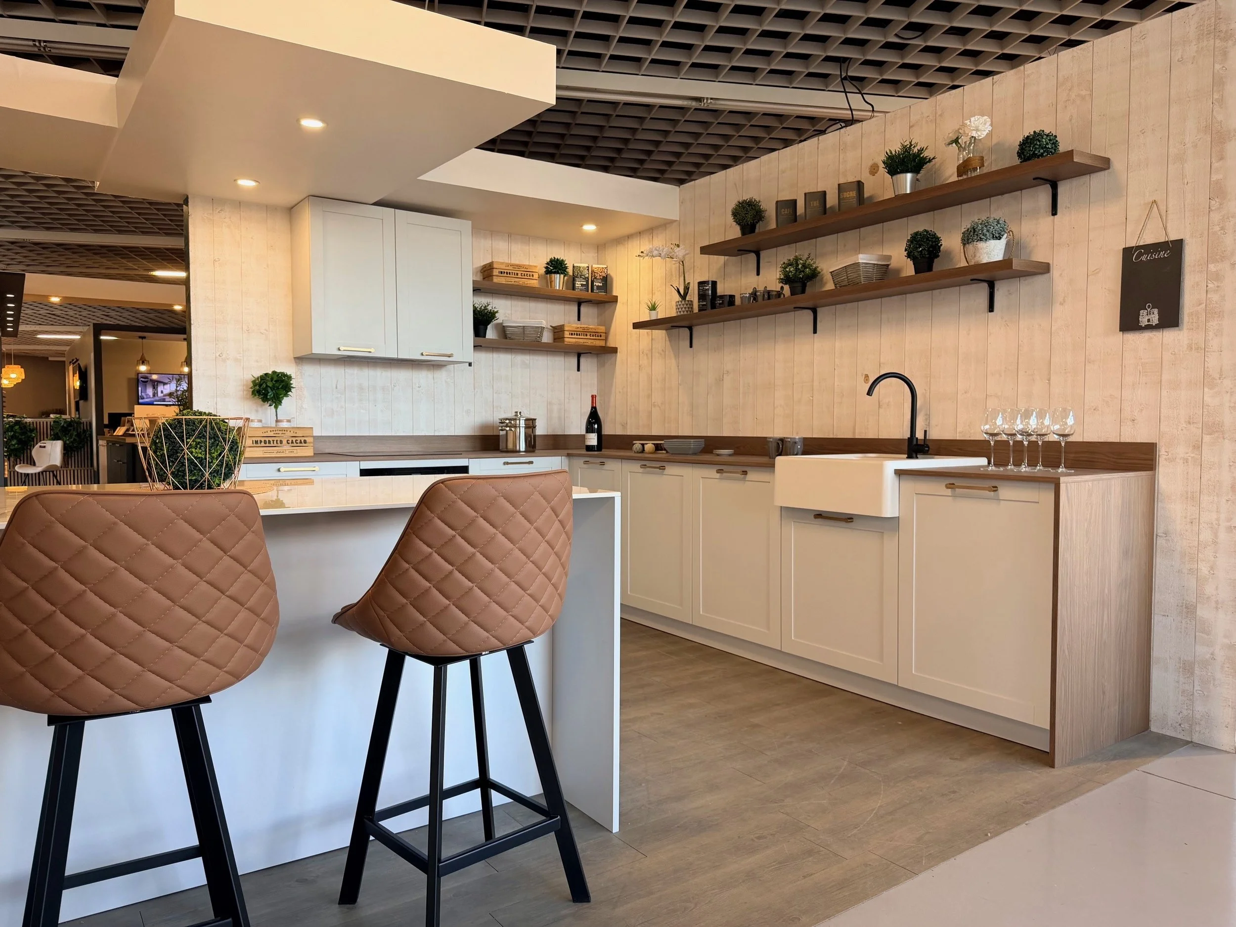 Cuisine moderne avec comptoir en bois, paniers, étagères avec plantes et objets décoratifs, deux chaises en cuir au premier plan.