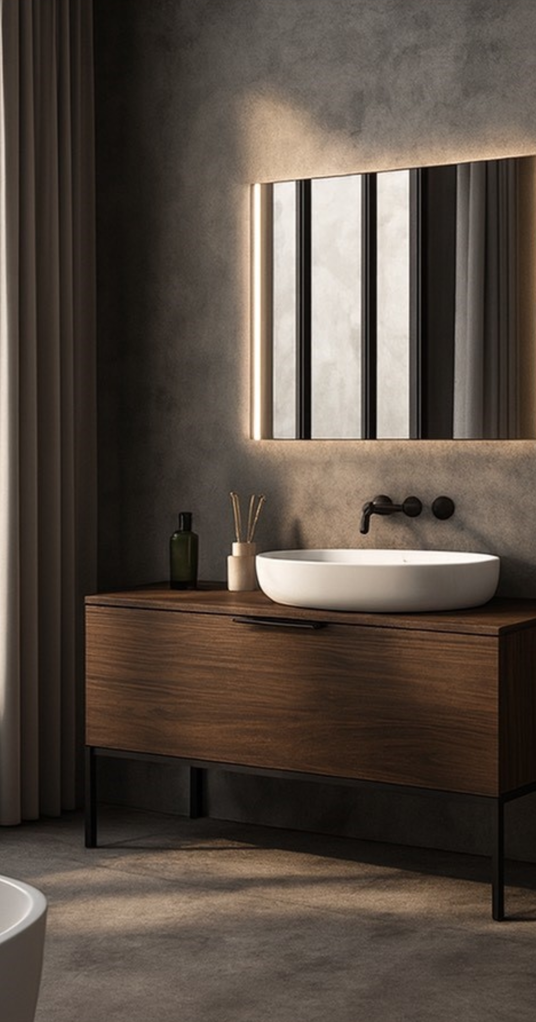 Salle de bain moderne avec un miroir rectangulaire entouré d'éclairage, un lavabo rond blanc sur une console en bois, un miroir avec grille en métal, une bouteille verte et un pot avec des bâtons de bois, rideau beige à gauche.