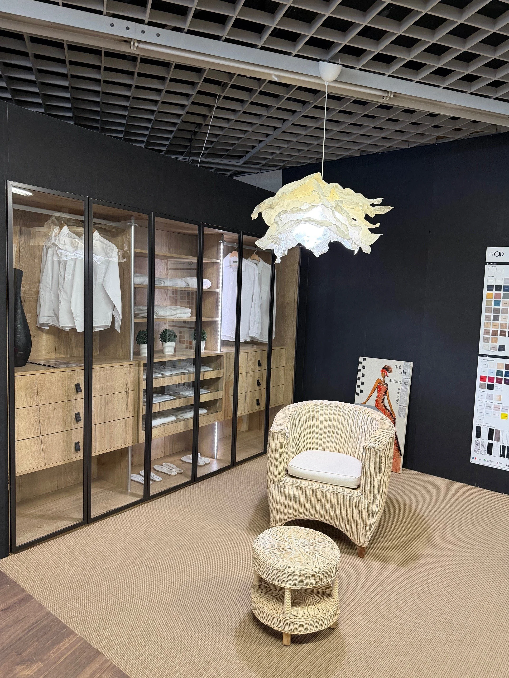 Une pièce décorative avec un fauteuil en rotin, un tabouret assorti, un luminaire en papier suspendu et une armoire en bois avec des vêtements blancs à l'intérieur, dans un espace contemporain.