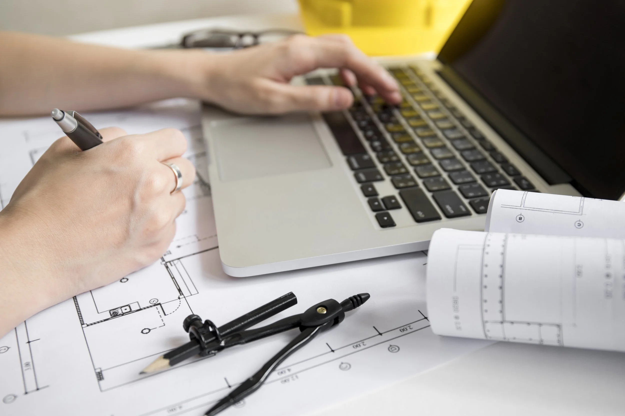 Une personne utilise un ordinateur portable avec des plans d'architecture, un compas, un crayon et des rouleaux de plans sur un bureau.