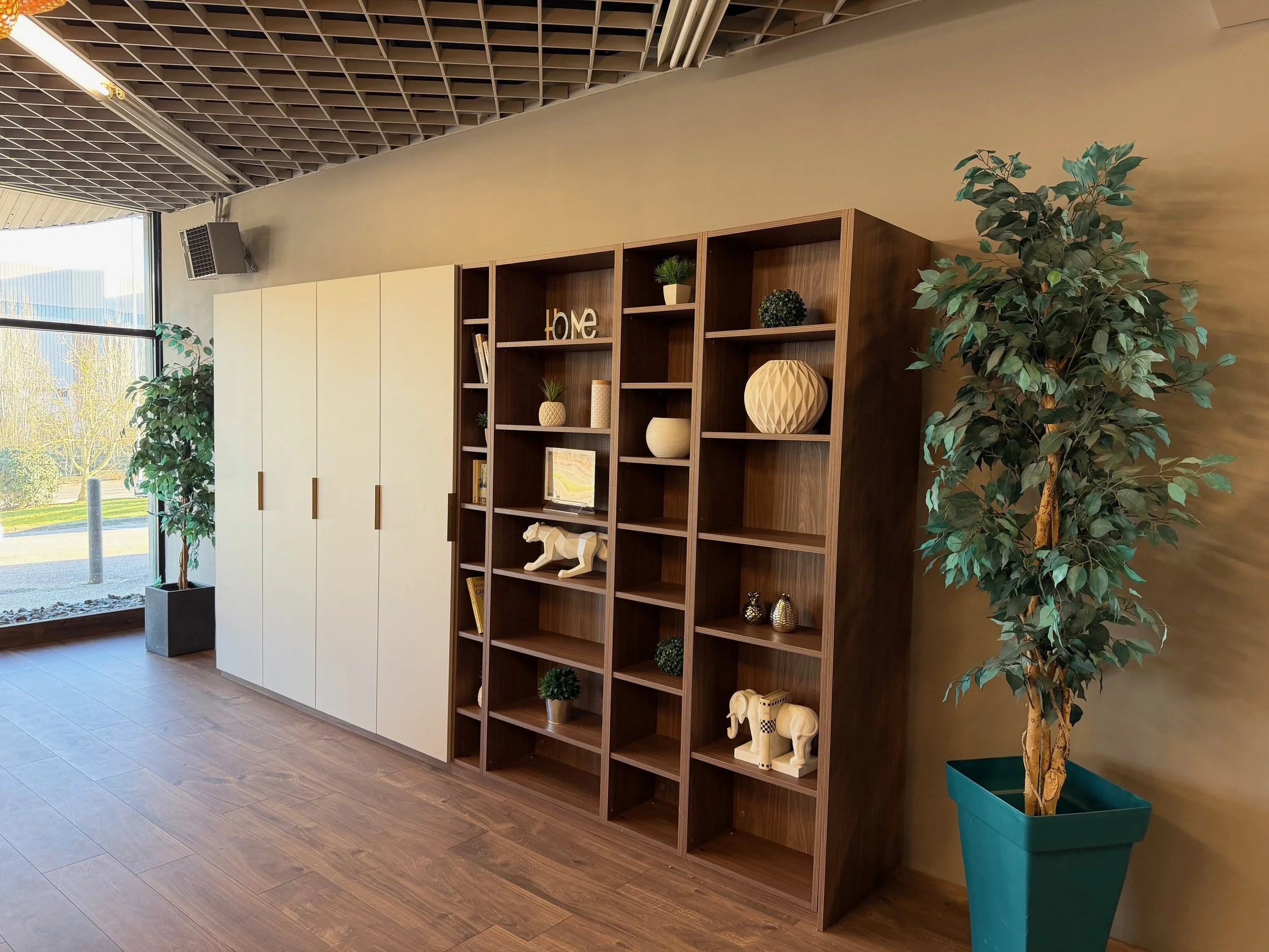 Une pièce avec de grandes fenêtres, un meuble en bois avec des étagères décoratives contenant des objets comme des plantes, des figurines d'éléphants, et des livres, ainsi que deux grands pots avec des plantes d'intérieur.