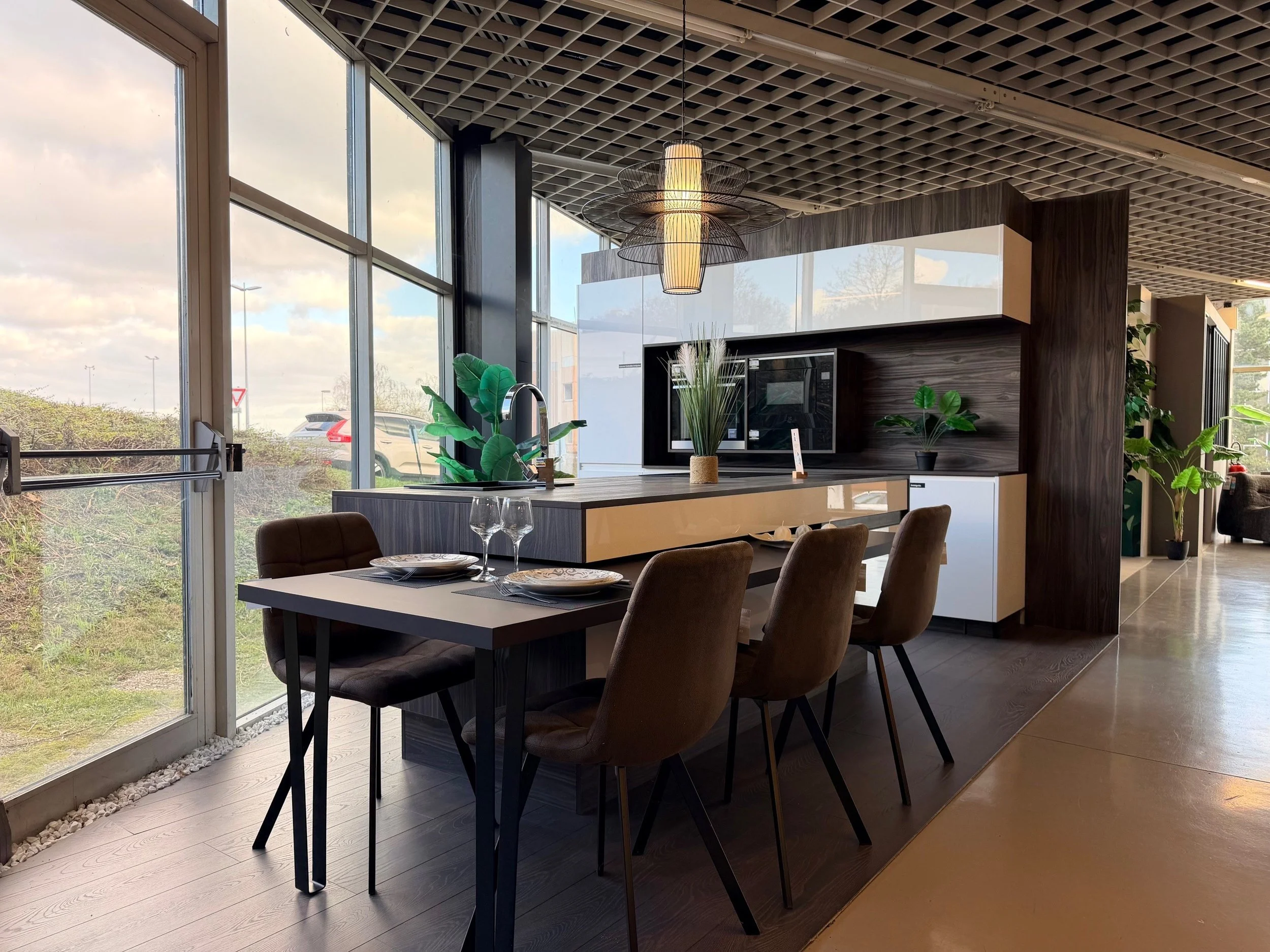 Cuisine moderne avec grand mur en bois foncé, comptoir en blanc, plusieurs plantes vertes, table à manger avec quatre chaises marron, repas prêt à être servi, luminaires suspendus au plafond, grandes fenêtres laissant entrer la lumière naturelle.