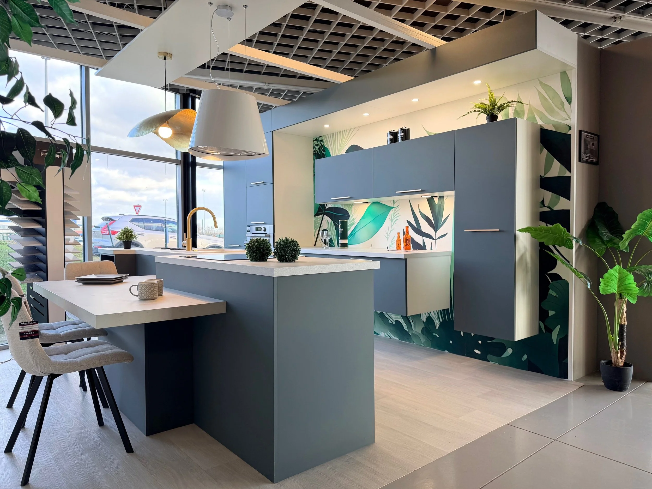 Cuisine moderne à thème végétal avec placards bleus, plan de travail blanc, mur en arrière-plan avec décoration de feuilles, grande fenêtre laissant entrer la lumière, plantes vertes, chaises confortables, vaisselle sur la table, éclairage suspendu.