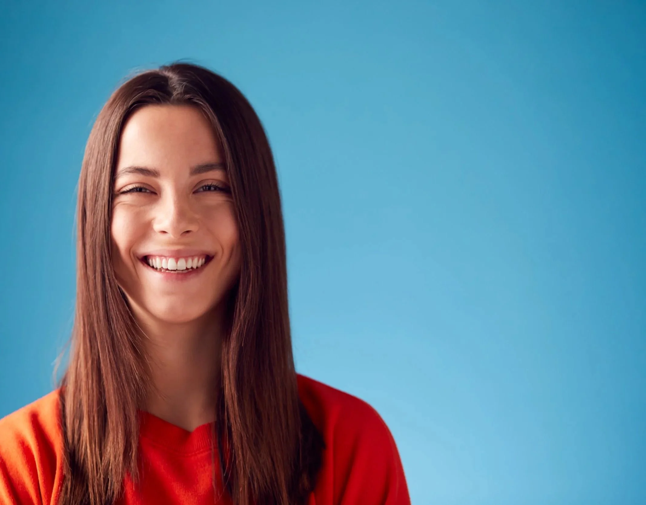 Young woman with long brown hair smiling against a bright blue background, wearing a red top.