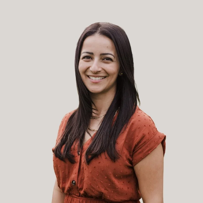 A woman with long dark hair smiling, wearing a rust-colored dress with short sleeves and black buttons, standing against a light gray background.