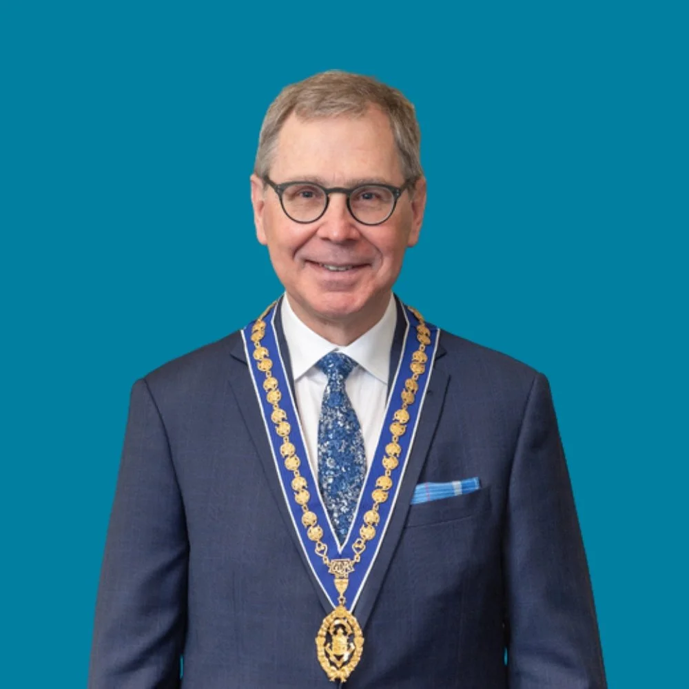 A middle-aged man wearing glasses, a dark suit, a patterned blue tie, and a white shirt. He has a blue and gold ceremonial chain around his neck and a matching pocket square, standing against a solid blue background.