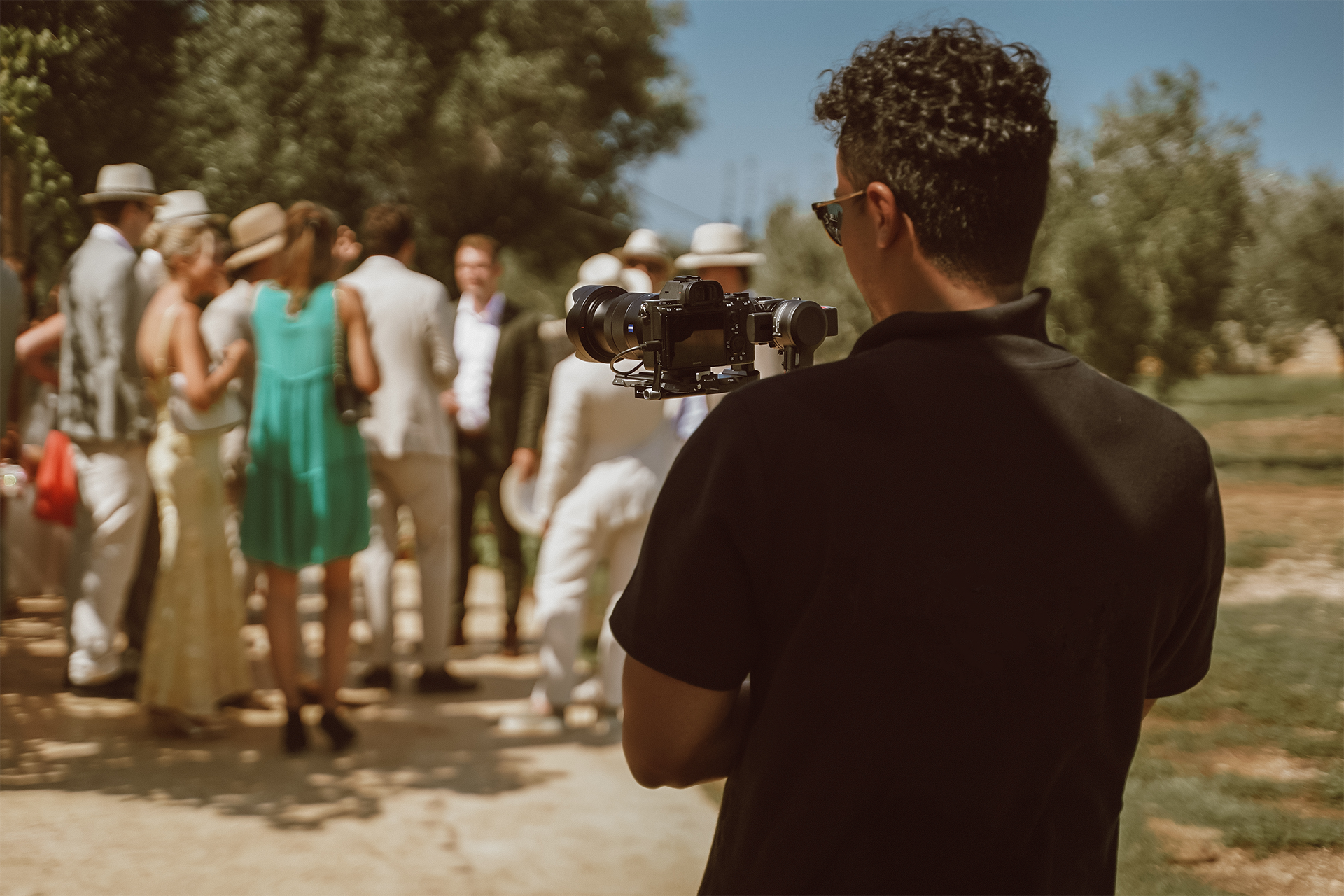 A person holds a camera and films a group of people outdoors during daytime, with trees in the background.