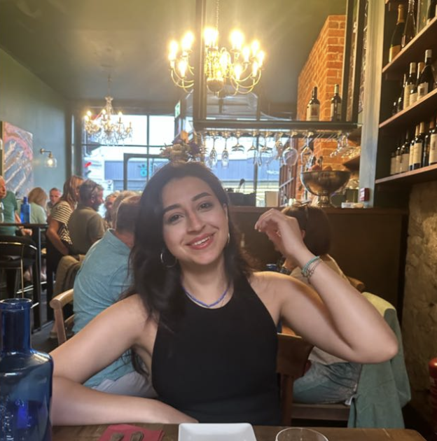 A young woman with dark hair and hoop earrings smiling at a restaurant, with a chandelier, brick wall, and other diners in the background.