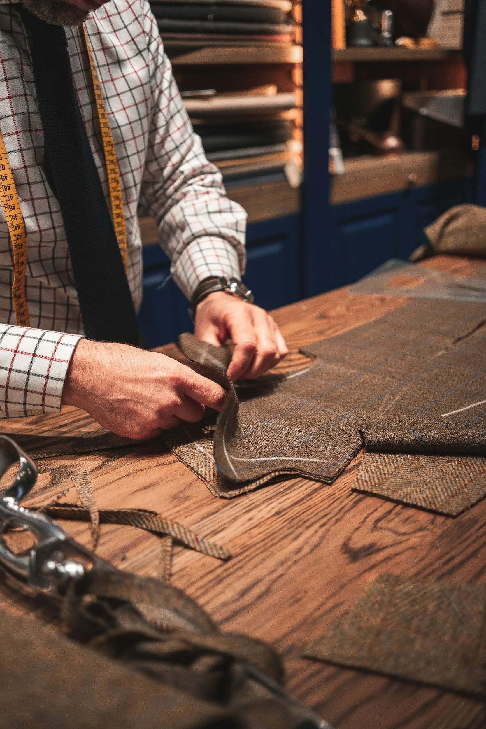 Person measuring and cutting fabric on a wooden worktable in a workshop, with shelves of tools and materials in the background.