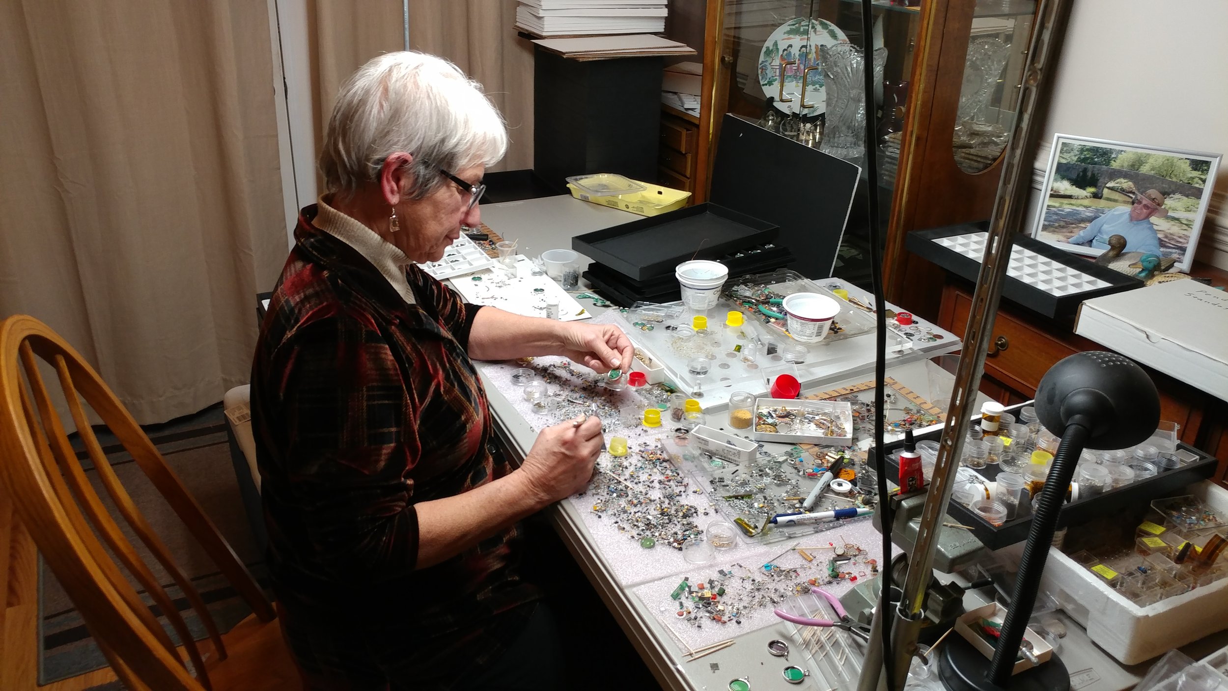 An elderly woman working on jewelry at a cluttered work table filled with beads, tools, and jewelry supplies, with a framed photo and display cases in the background.