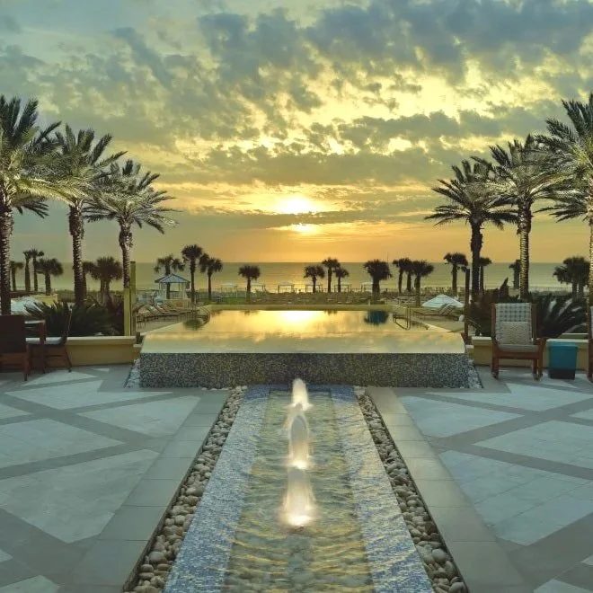 Beautiful sunset over a luxurious pool area at the Omni Resort Amelia Island with water fountains, surrounded by palm trees and outdoor seating, overlooking the ocean.