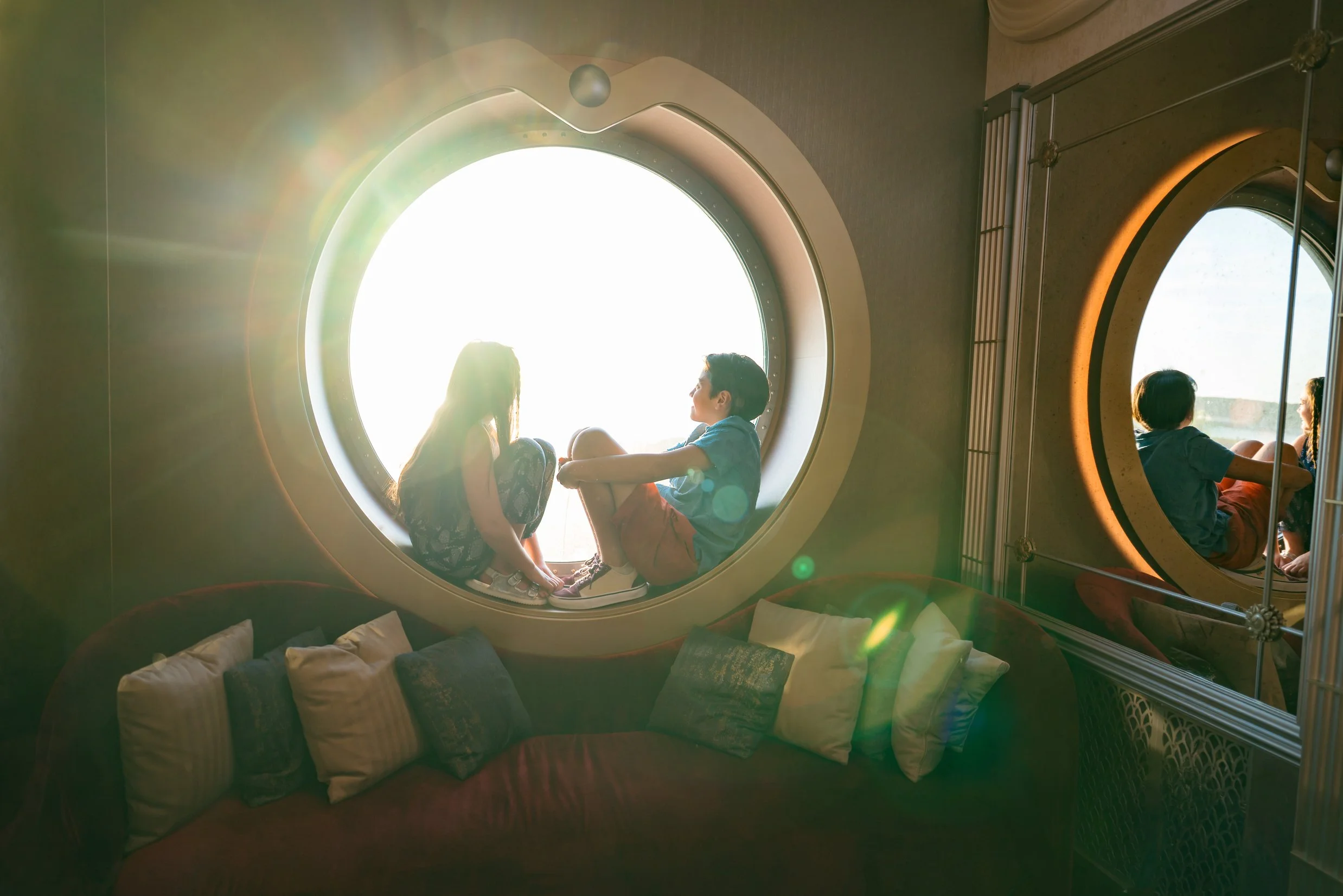Two children sitting together in a circular window seat, backlit by bright sunlight, with a reflection of them and another person in a mirror on the right.