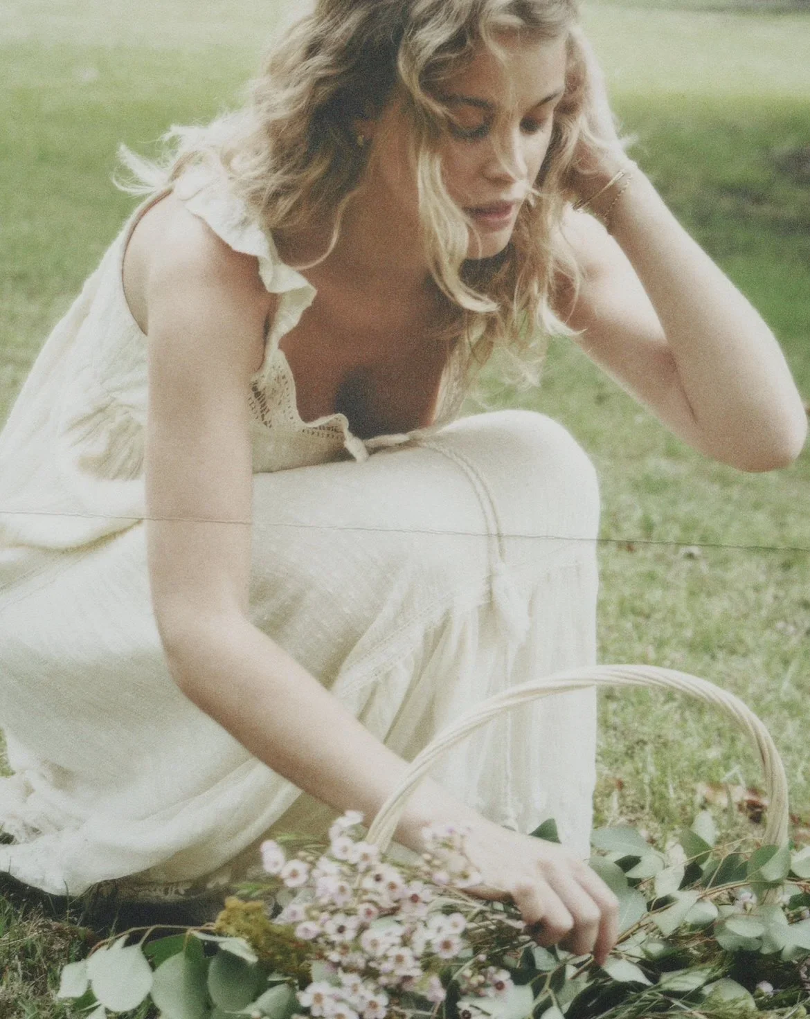 A woman in a white dress sitting on the grass, holding a basket of pink flowers and greenery.