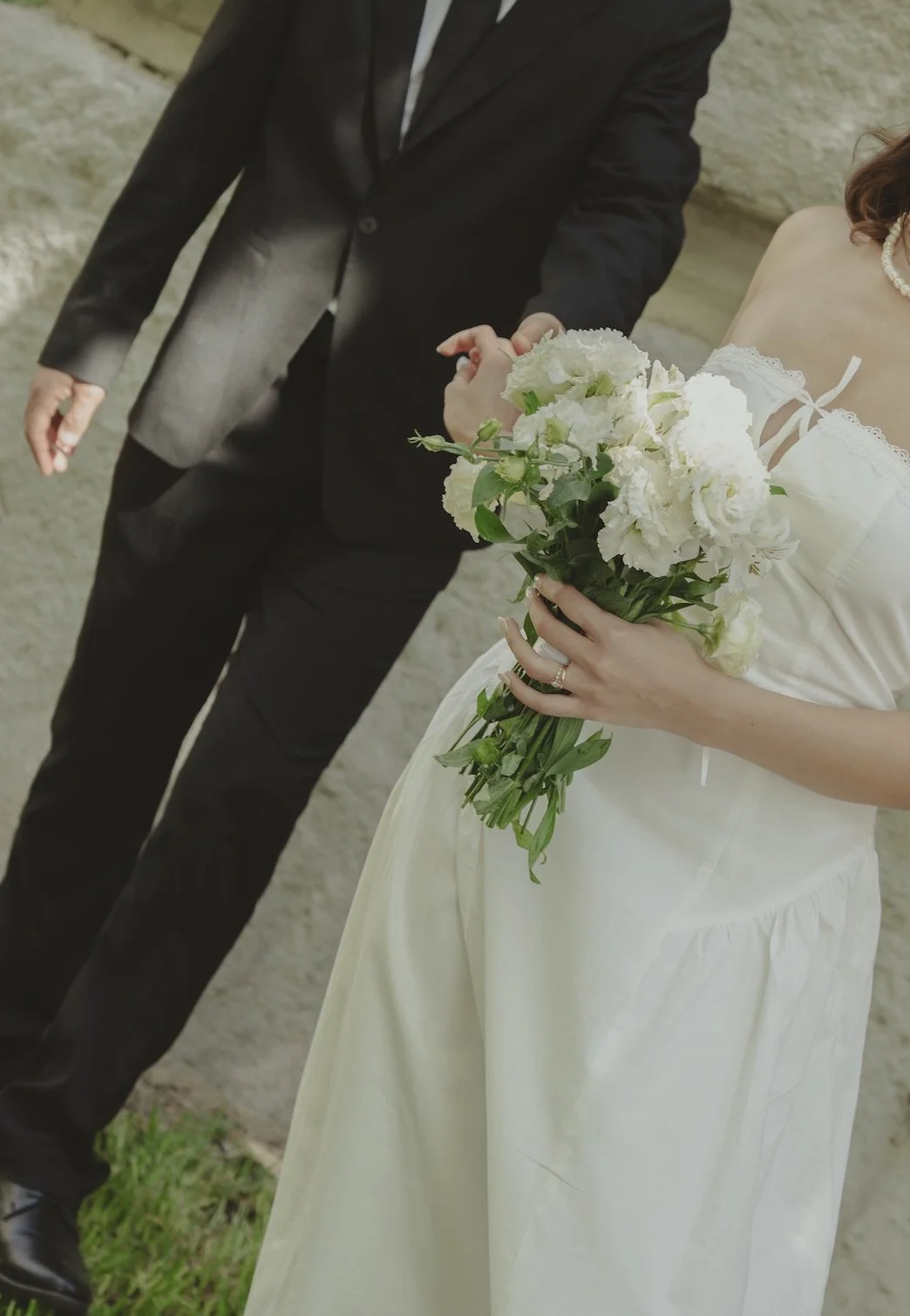 A bride in a white wedding dress holding a bouquet of white flowers, with a groom in a black suit standing next to her.
