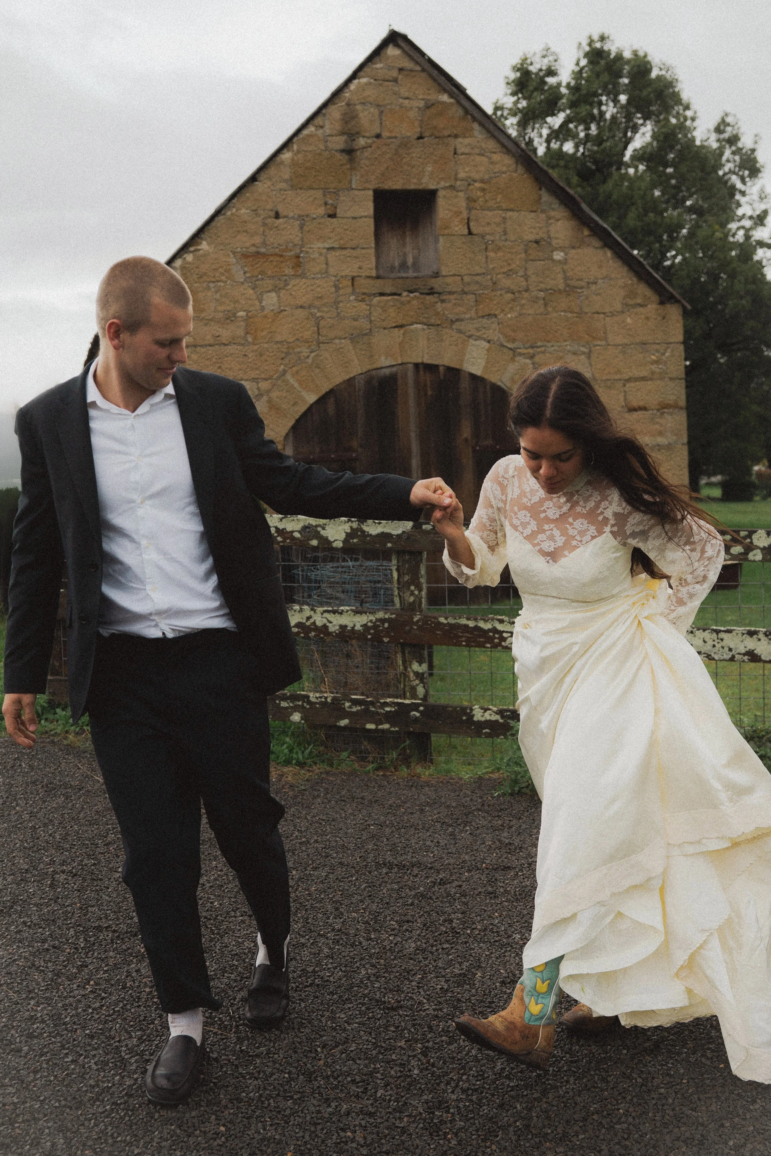 A couple walking hand in hand near a rustic barn, with the woman wearing a wedding dress and cowboy boots, and the man in a black suit and white shirt.
