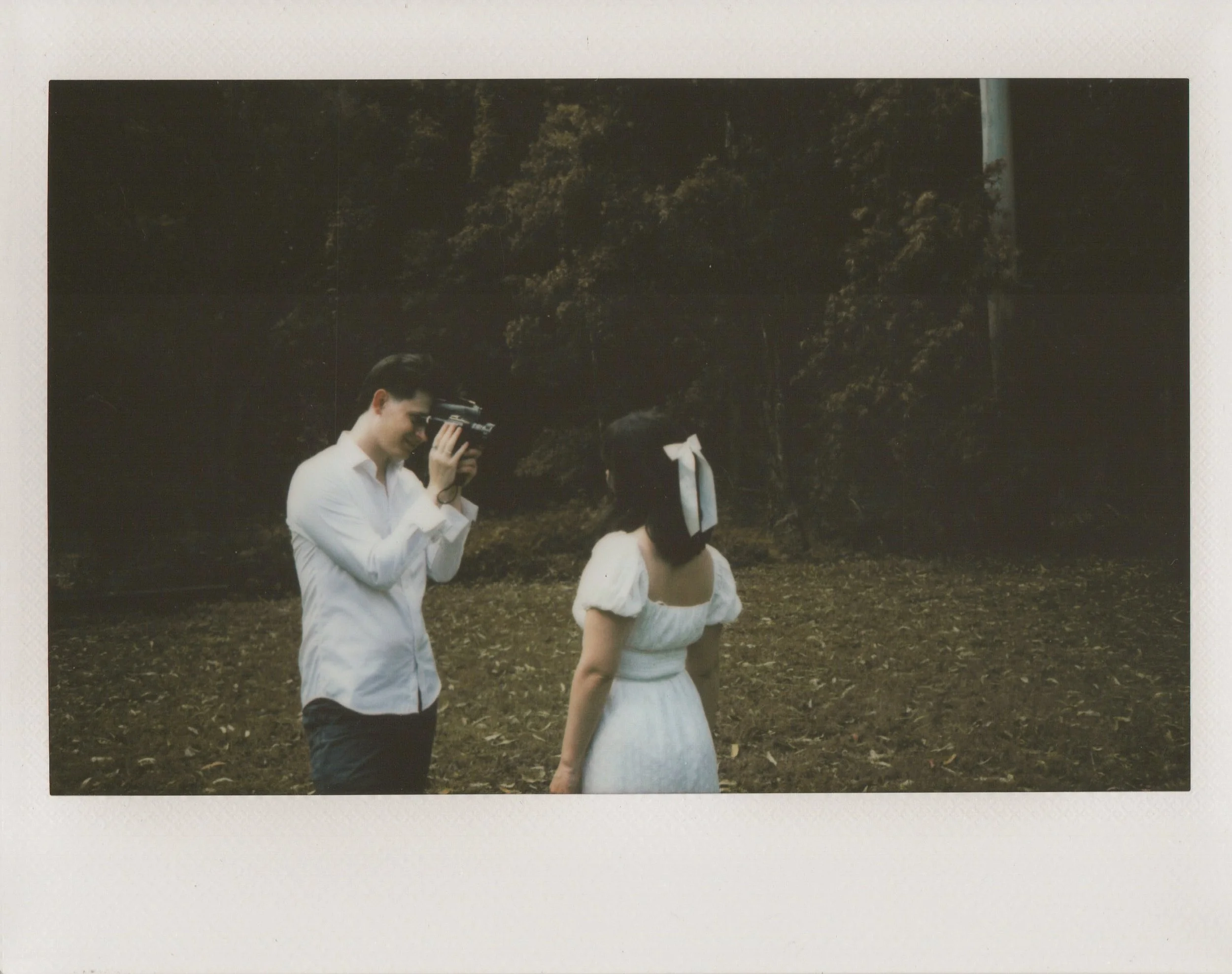 A man in a white shirt is taking a photograph of a woman in a white dress with a ribbon in her hair in a forested area.