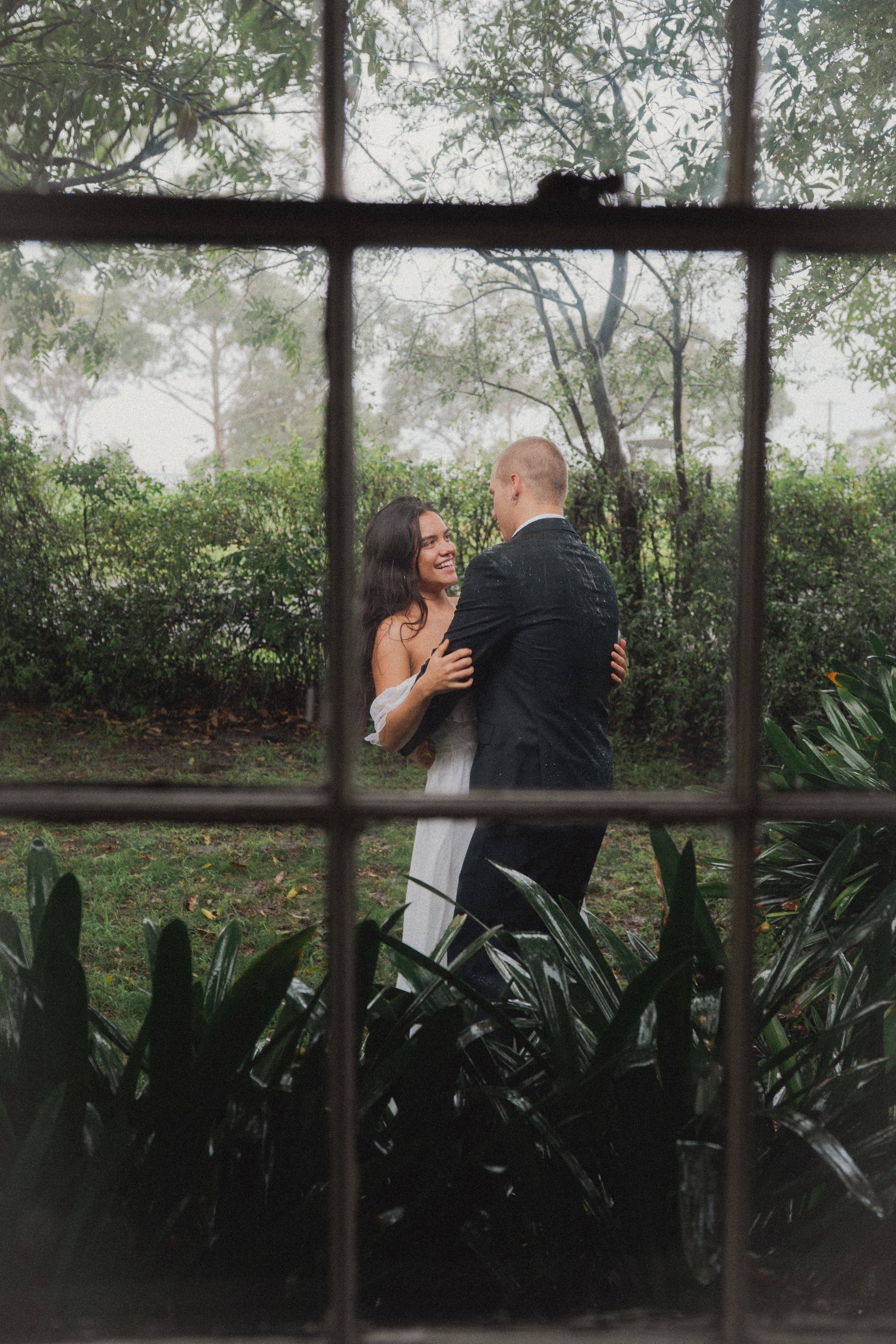 A bride and groom dancing outdoors on a foggy day, seen through a window with grid panes. Wedding attire, trees, and plants are visible in the background.