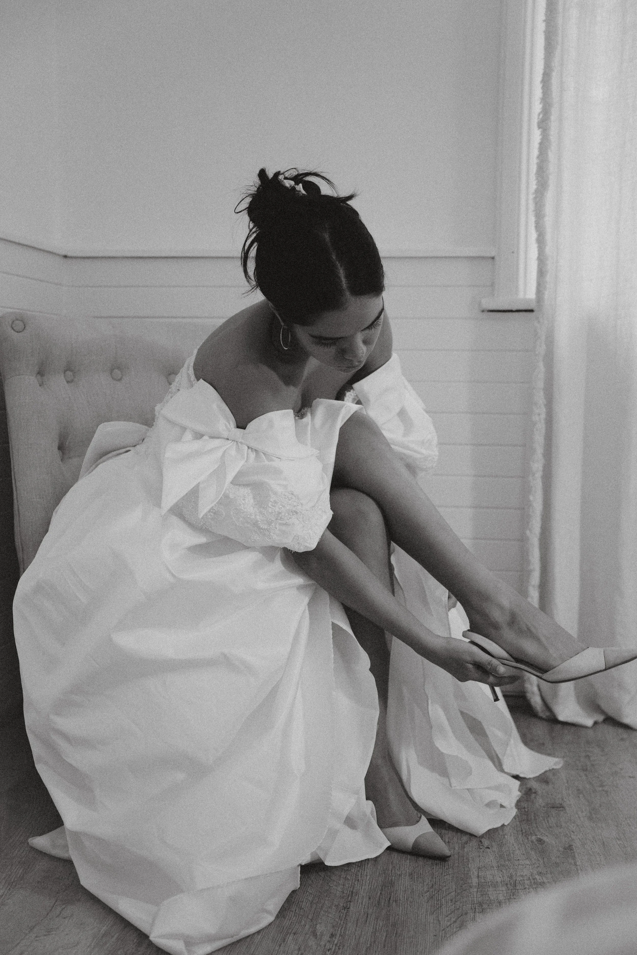 A woman in a wedding dress is sitting on a cushioned chair, putting on her high-heeled shoe.