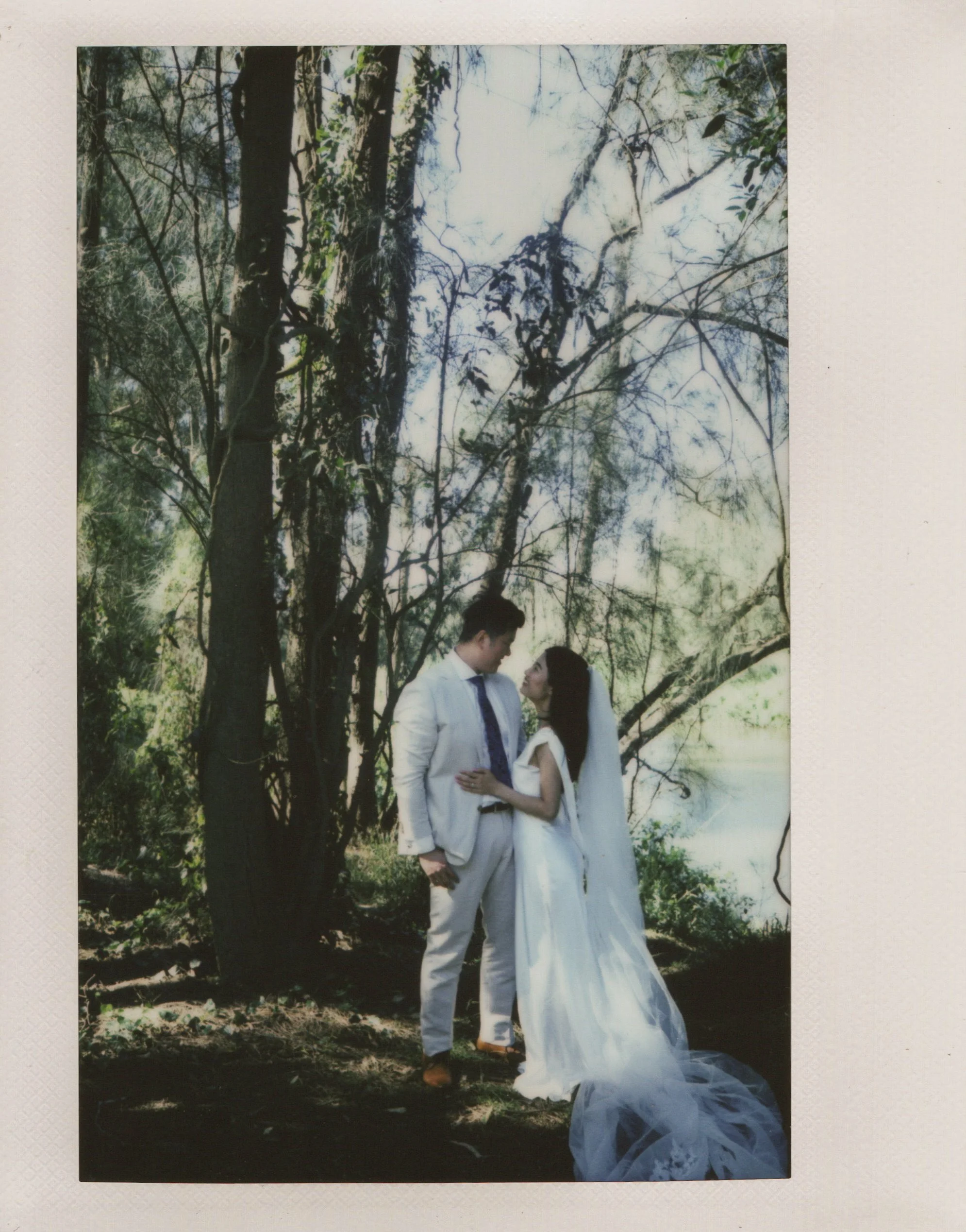 A bride and groom in wedding attire standing outdoors in a wooded area with trees and sunlight filtering through, looking into each other's eyes.