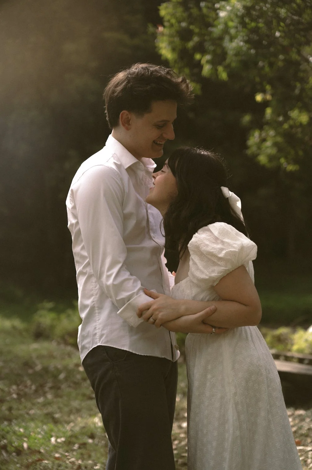 A couple in love standing outdoors, smiling, and gazing into each other's eyes, holding hands, surrounded by greenery.