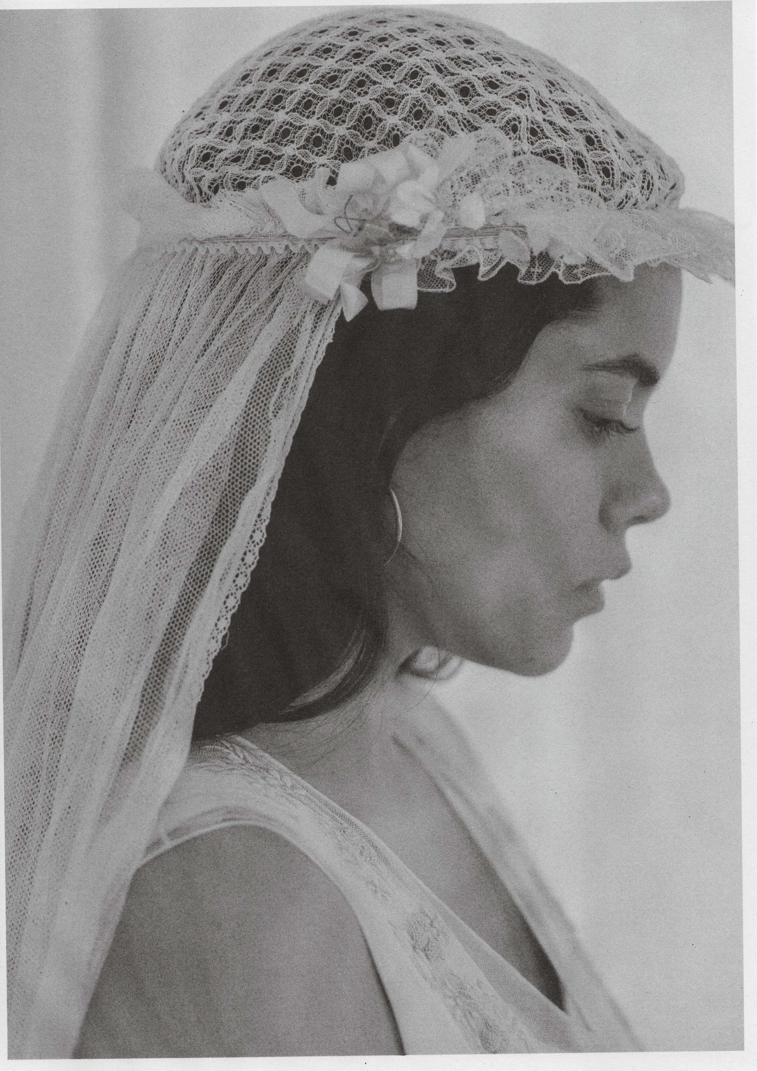 A black and white photo of a woman in profile, wearing a lace cap with a veil and a flower decoration, with her eyes closed and a calm expression.