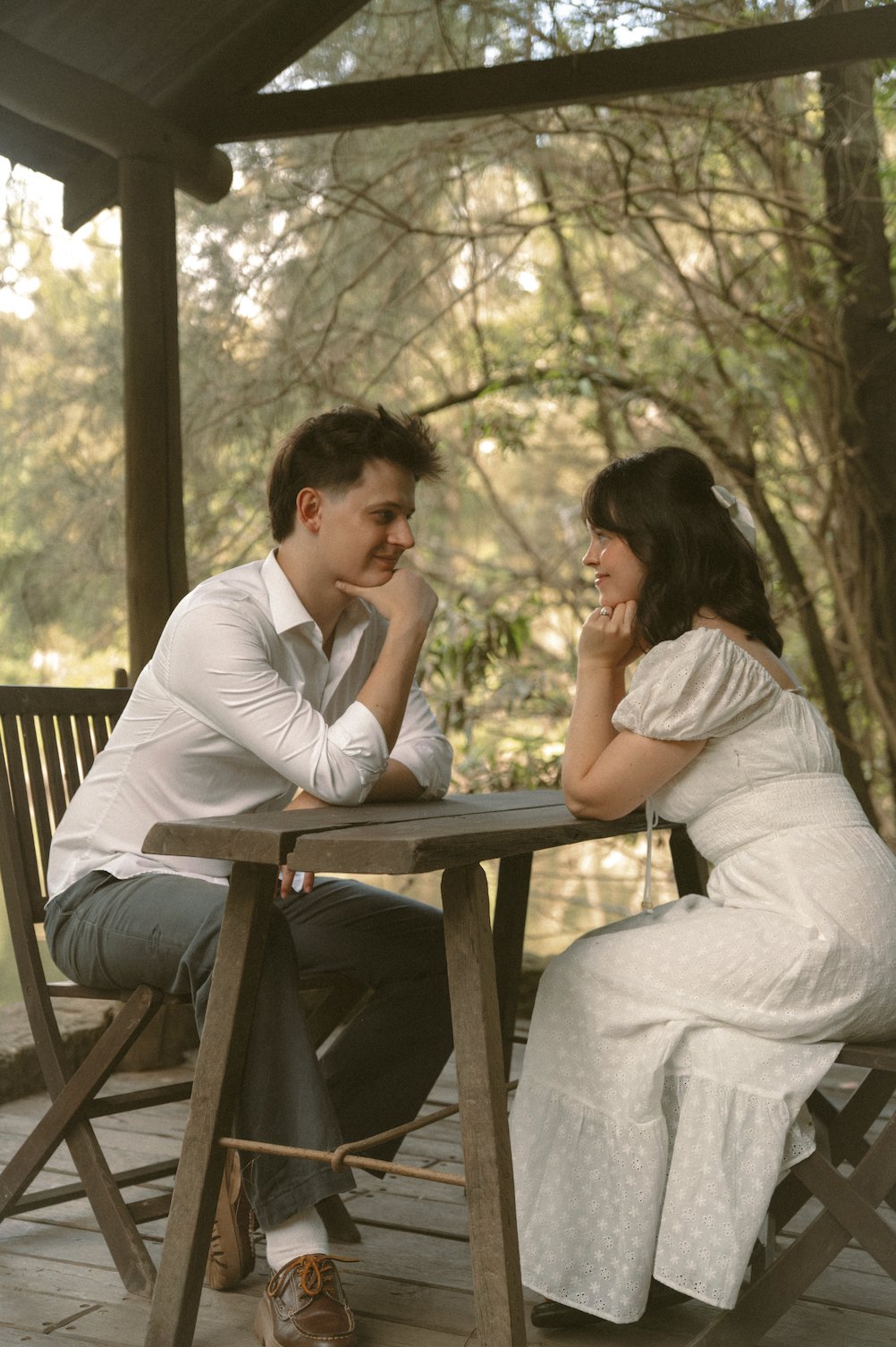 A young man and woman sit across from each other at a wooden table on an outdoor porch, gazing into each other's eyes with warm, affectionate expressions, surrounded by trees and soft natural light.