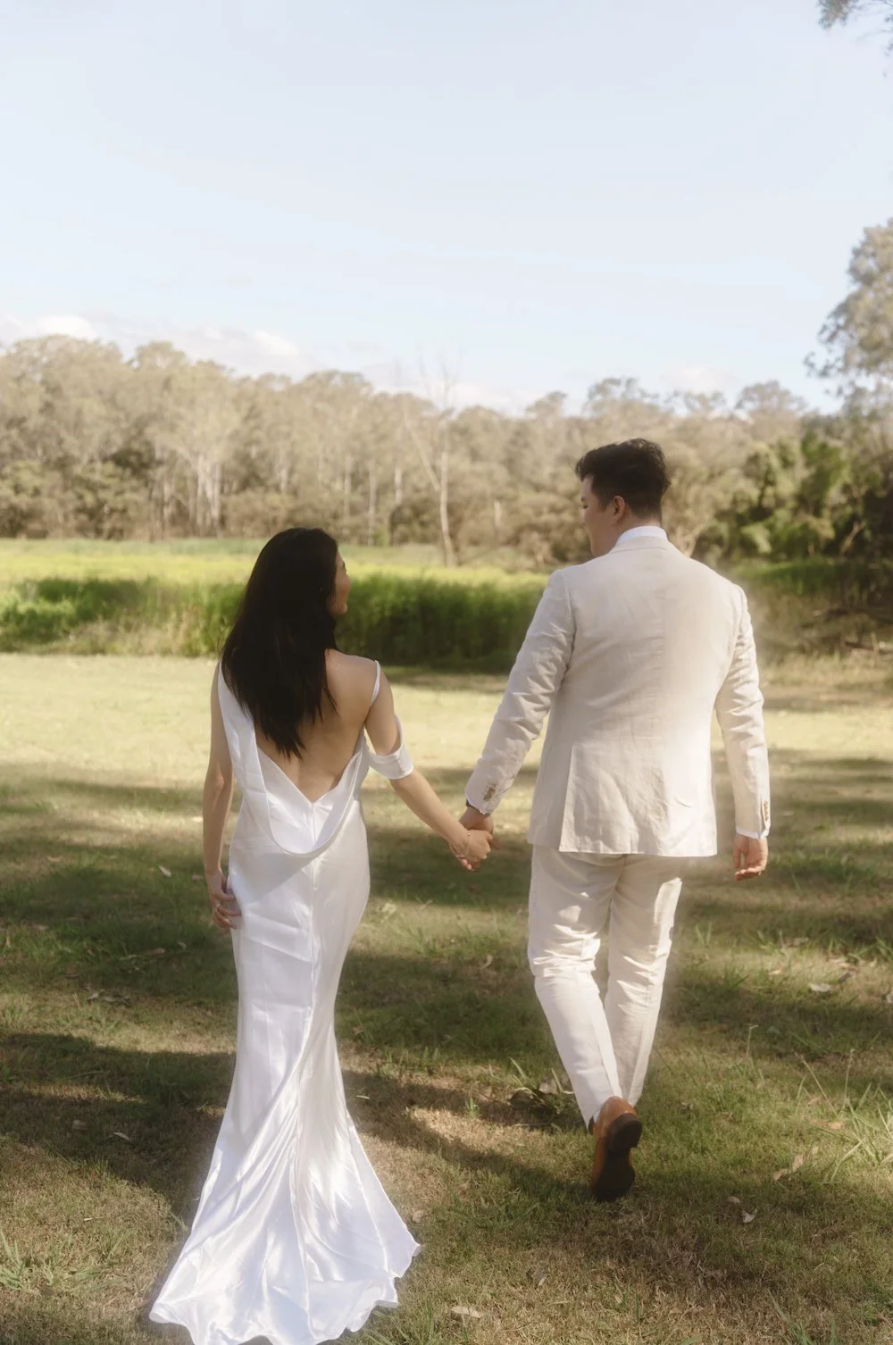 A couple in white wedding attire holding hands and walking in a grassy outdoor area with trees and a blue sky in the background.