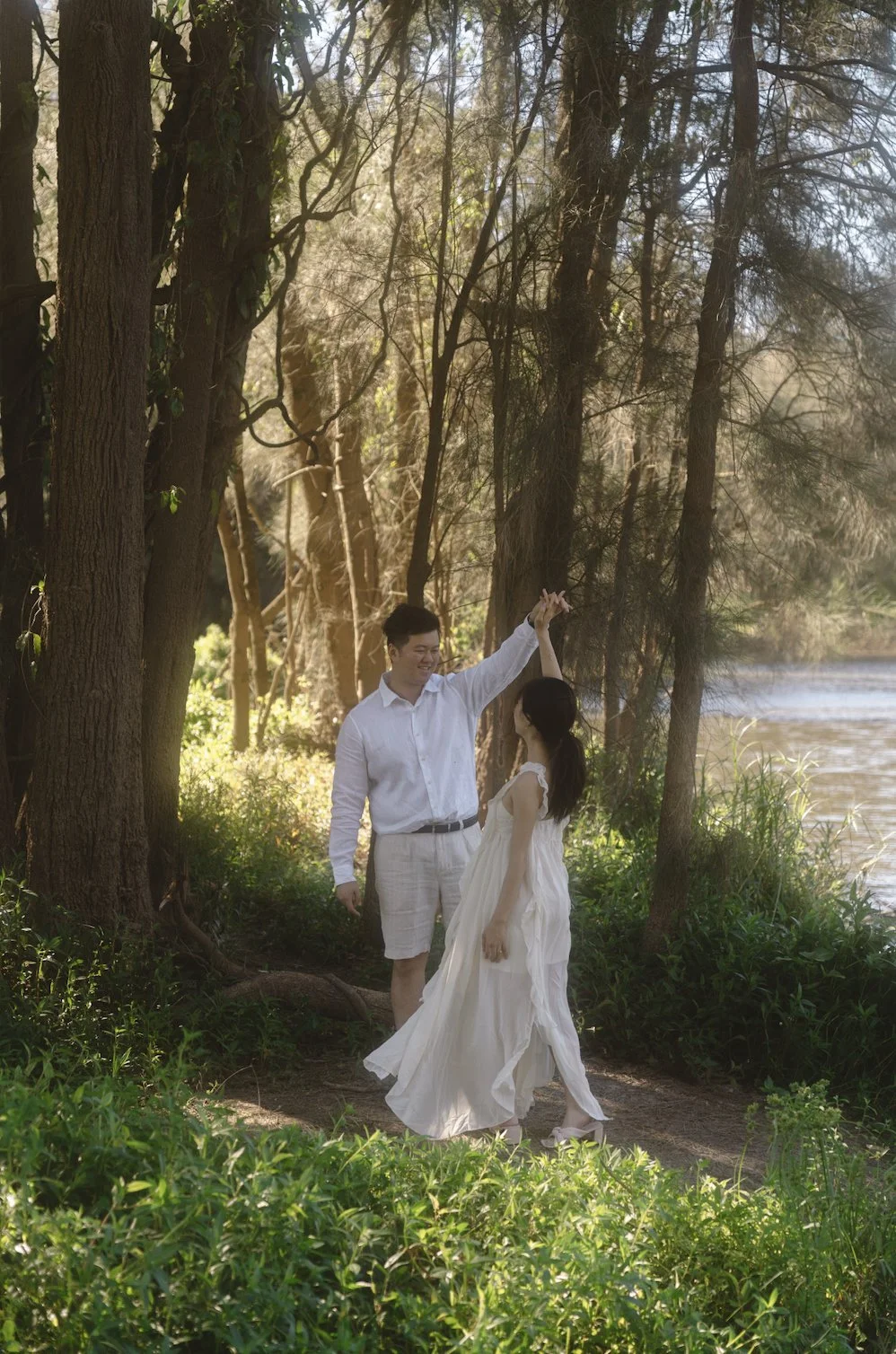 A young couple dancing and smiling under tall trees near a river, surrounded by lush greenery in a peaceful outdoor setting.