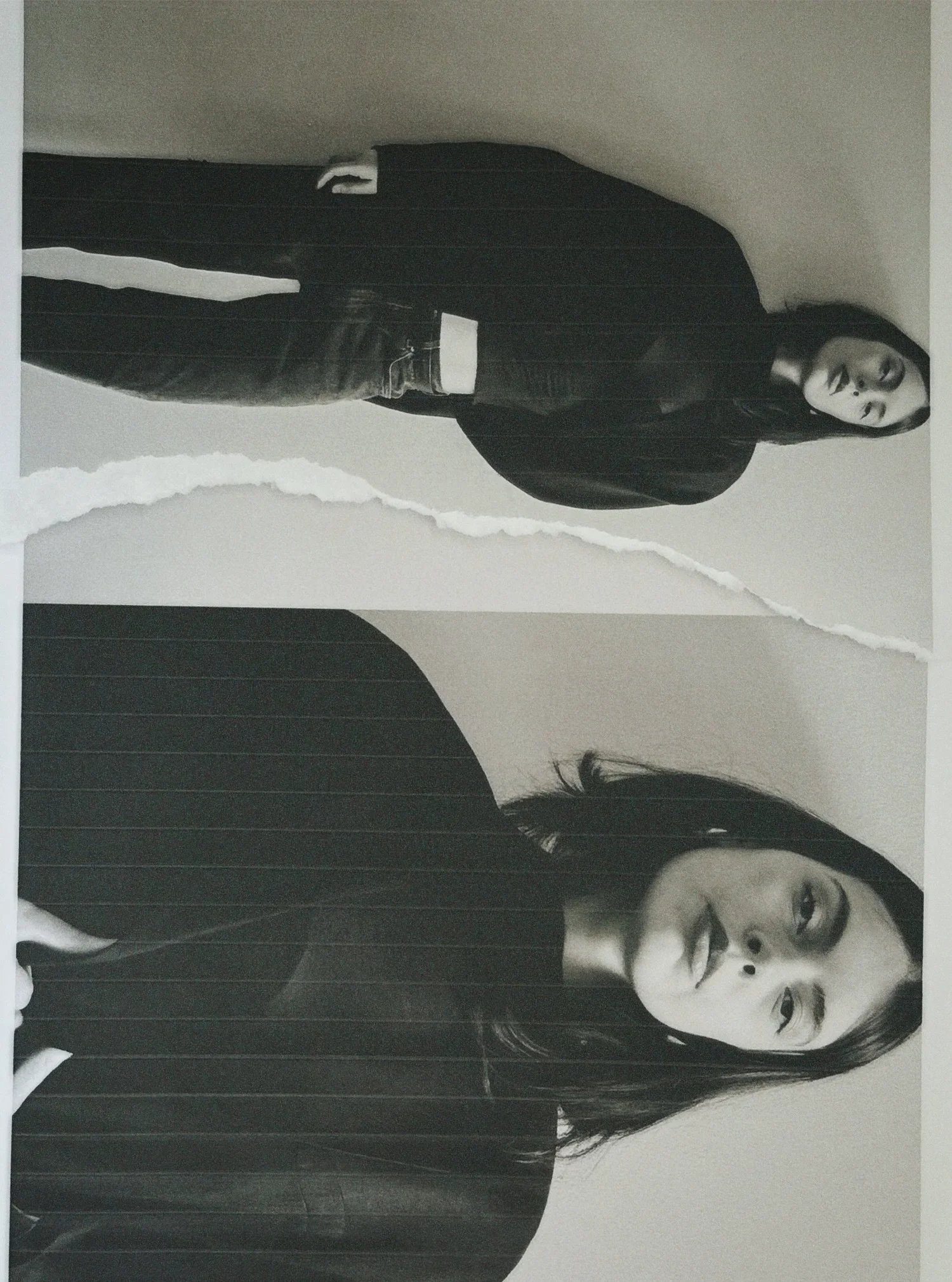 Black-and-white photograph of two women standing in front of an abstract, torn-paper background. The woman in the foreground has long dark hair, wears a dark striped shirt, and looks directly at the camera with a neutral expression. The woman in the 