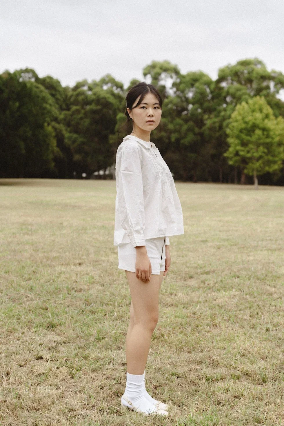 A young woman standing in an open grassy field with trees in the background, wearing a white blouse, white shorts, white socks, and sandals.
