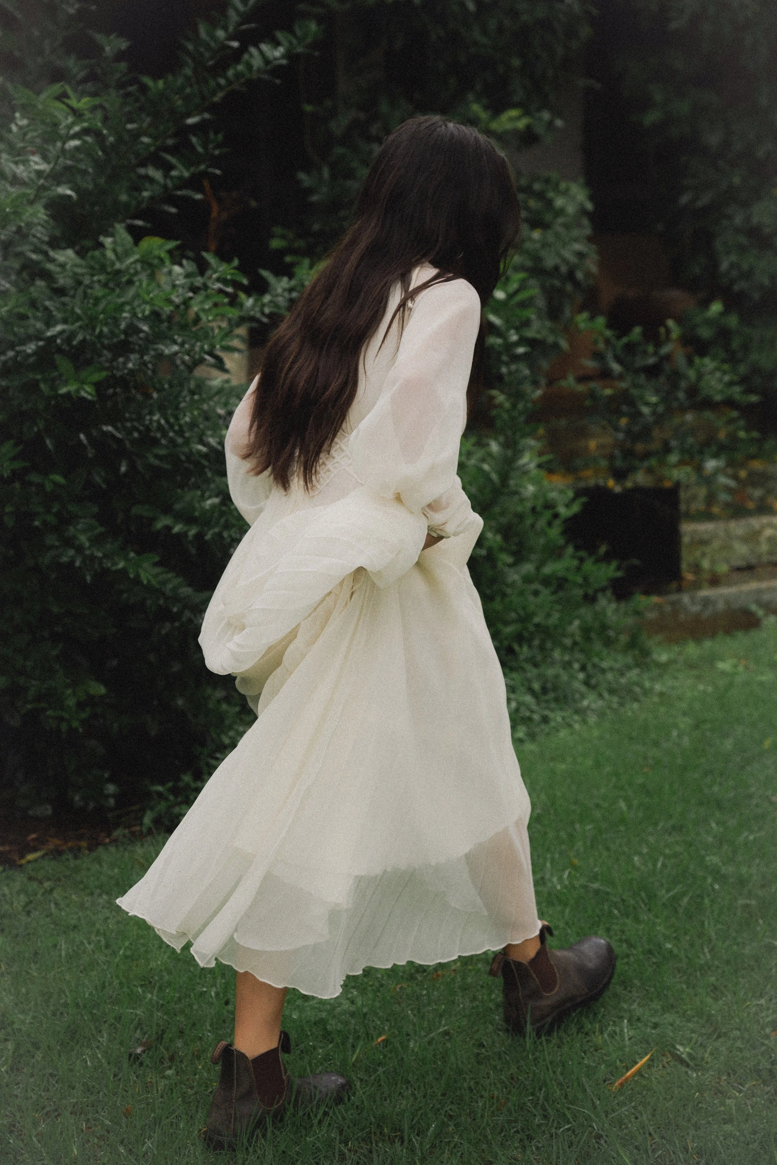 A woman with long dark hair wearing a white dress and dark boots walking through a garden with lush green foliage.