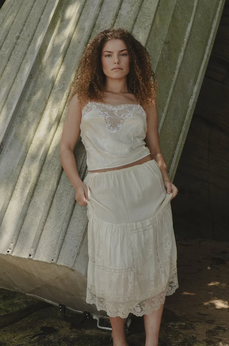 A woman with curly hair wearing a cream-colored camisole top with lace details and a matching long skirt, standing outdoors against a stone wall.