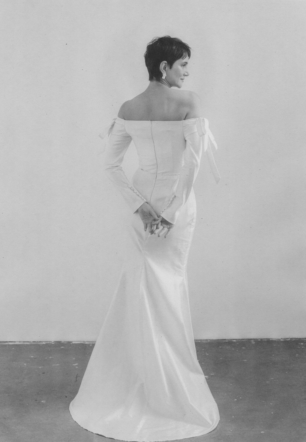 A woman with a short hairstyle wearing an elegant off-the-shoulder gown with long sleeves and a train, standing against a plain wall.