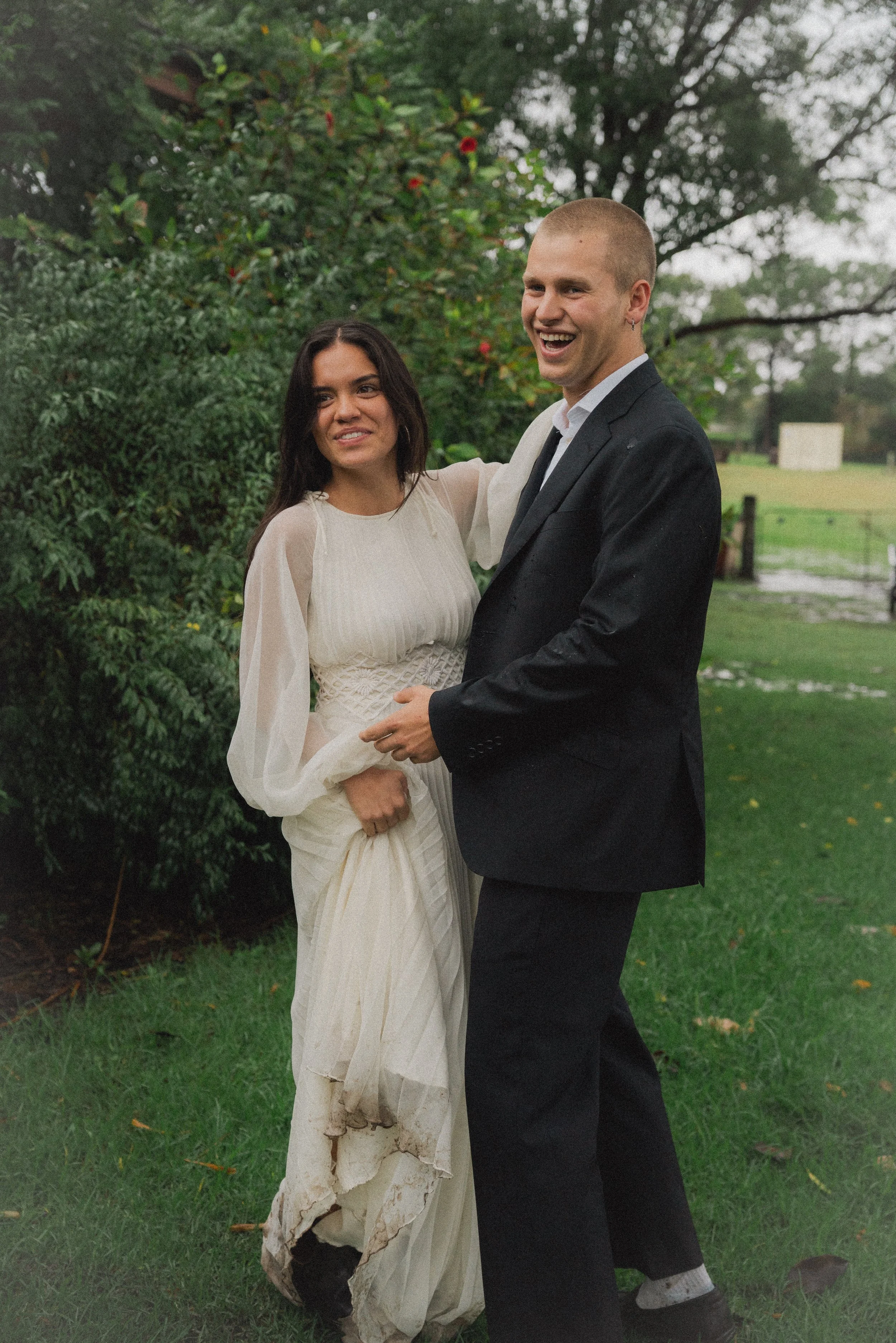 Minimalist photo of a couple in wedding attire outdoors on a rainy day, standing on grass with green bushes and trees in the background, the woman in a cream-colored dress with mud stains and the man in a black suit, both smiling.