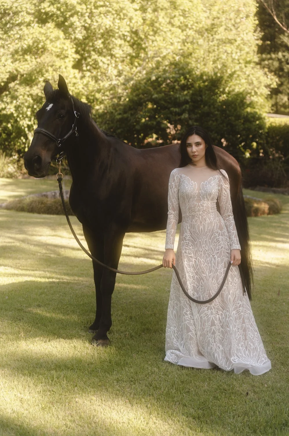 Woman in a long, white embroidered dress holding a black horse's lead in a lush, green outdoor setting.