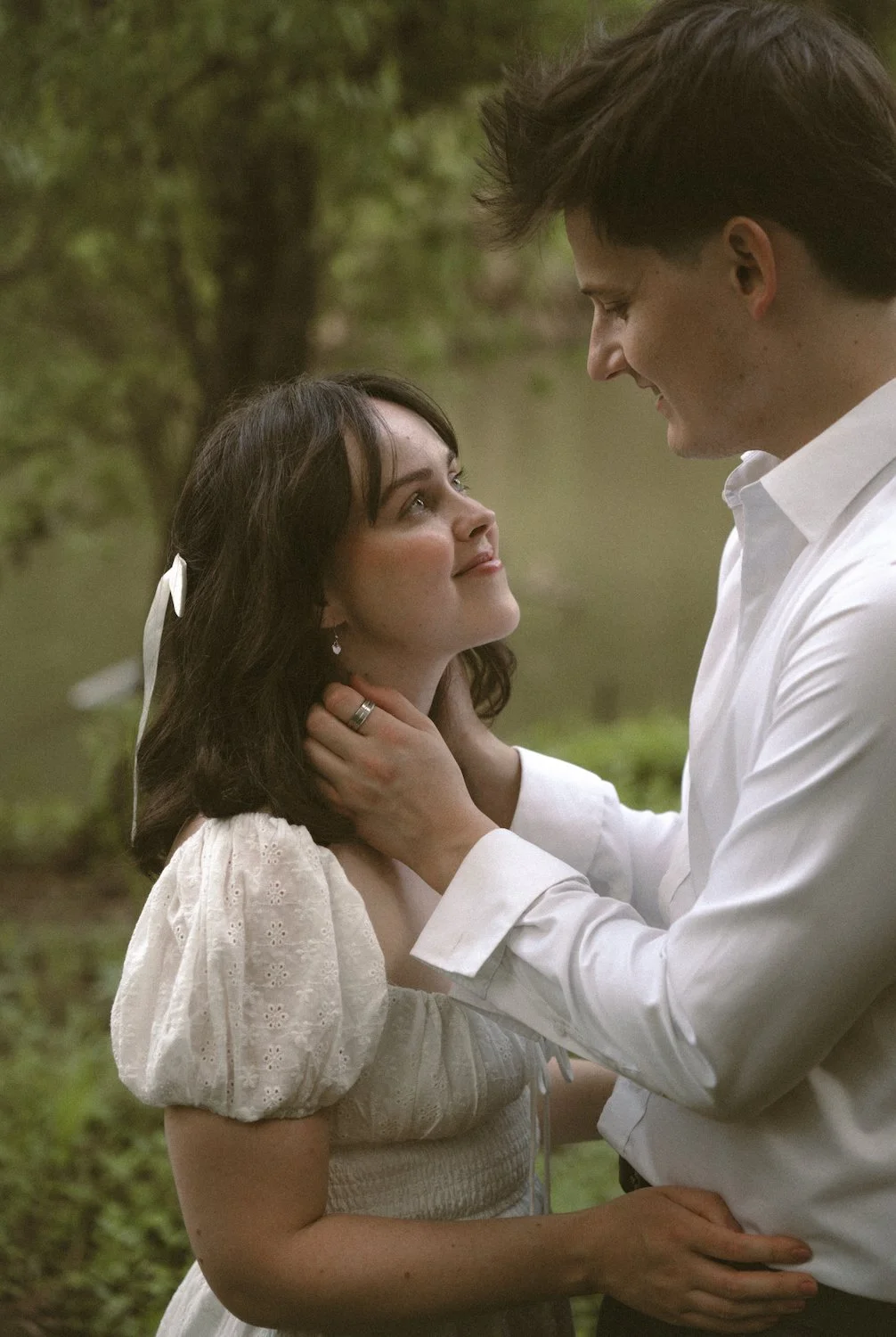 A man holding a woman's face tenderly, both smiling and looking into each other's eyes, outdoors near a body of water with green trees in the background.