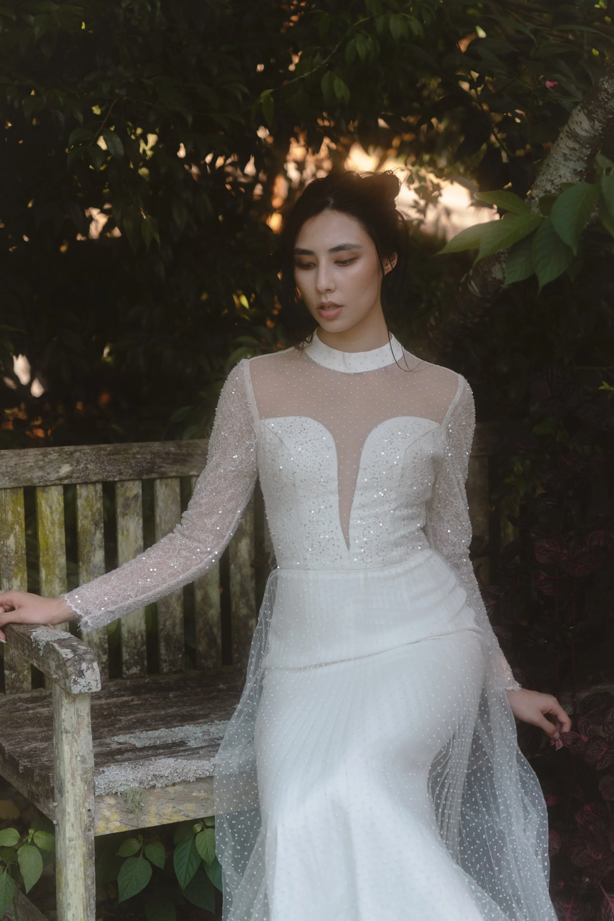 A woman in a white, long-sleeved, sparkly dress sitting on a weathered wooden bench outdoors with lush greenery in the background during sunset.