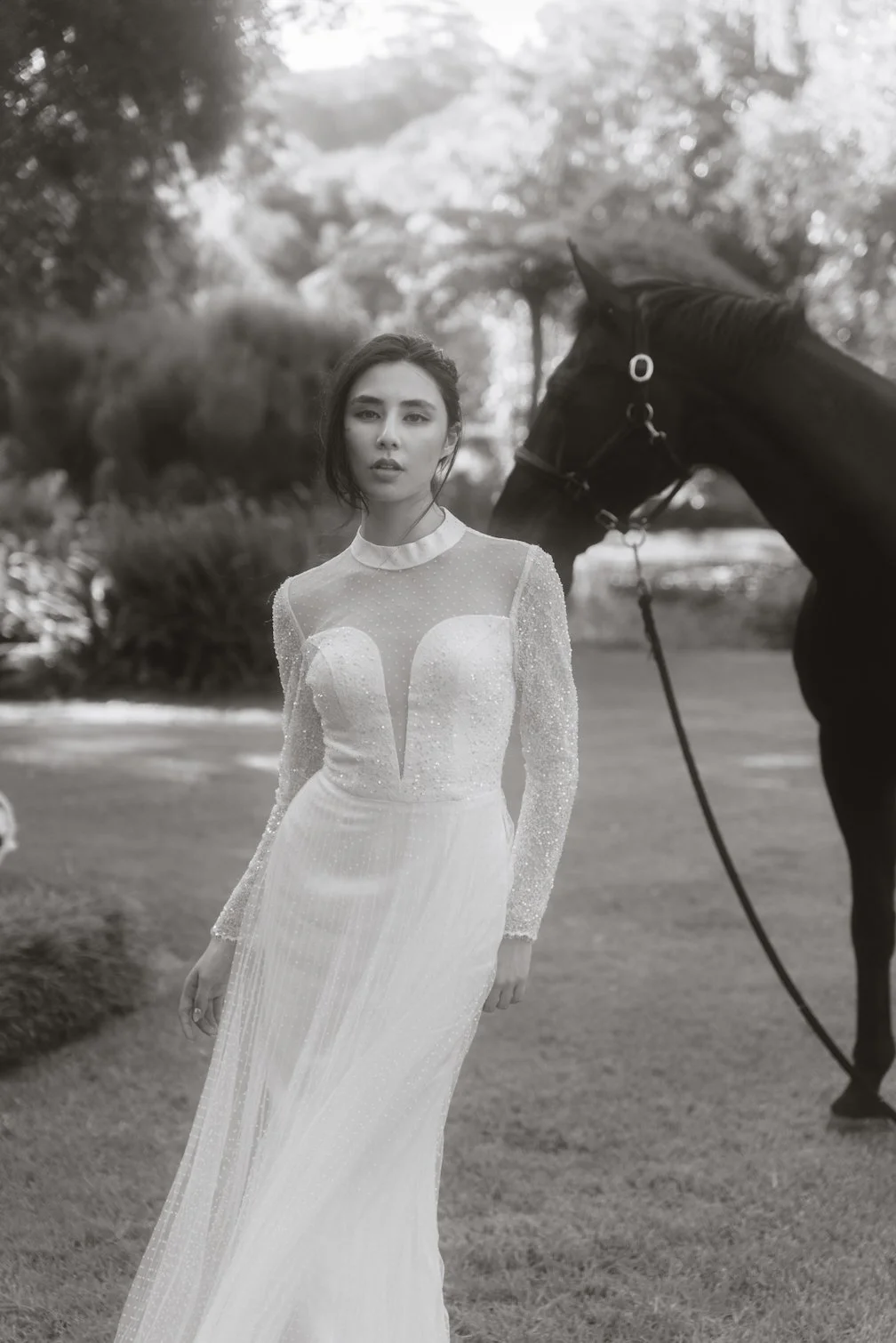 A woman in a white, embellished gown standing outdoors next to a black horse, with trees and grass in the background.
