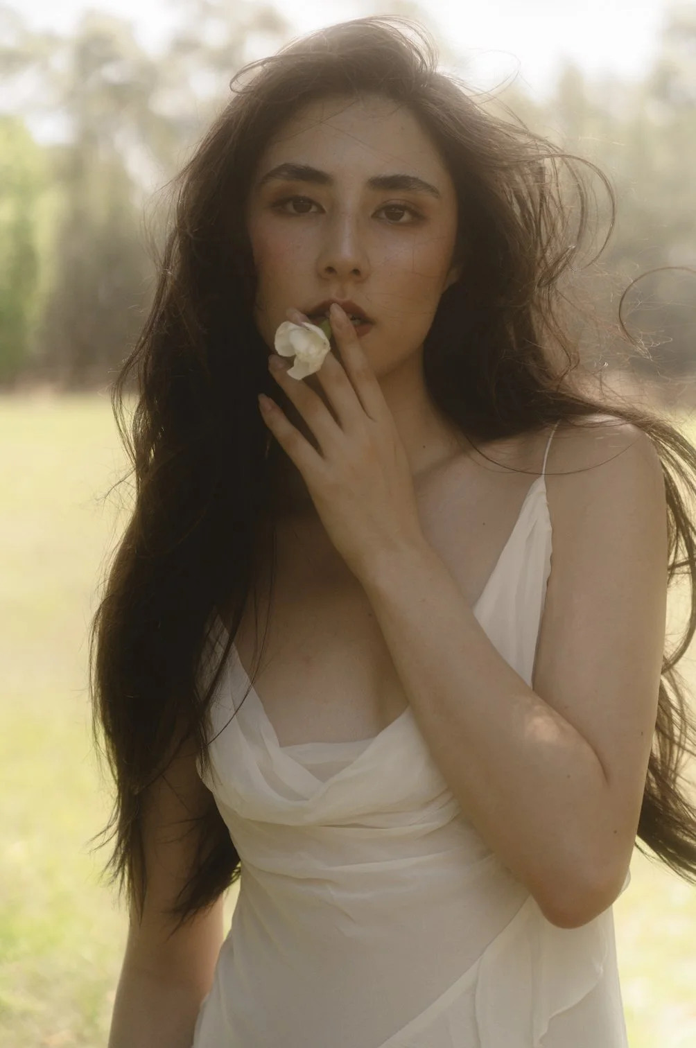 A woman with long dark hair and light skin, wearing a white dress, poses outdoors with a flower near her lips in front of a blurred background of trees and sunlight.