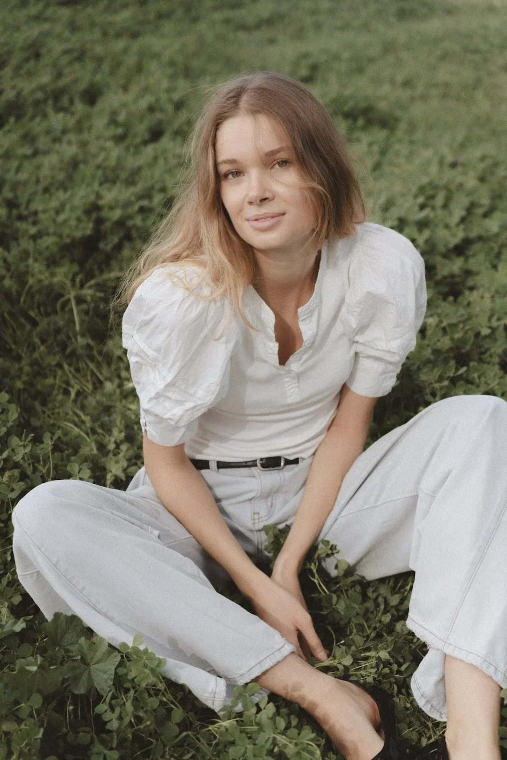 A young woman with long, wavy blonde hair sitting on the ground in a grassy area, wearing a white blouse and light-colored pants.