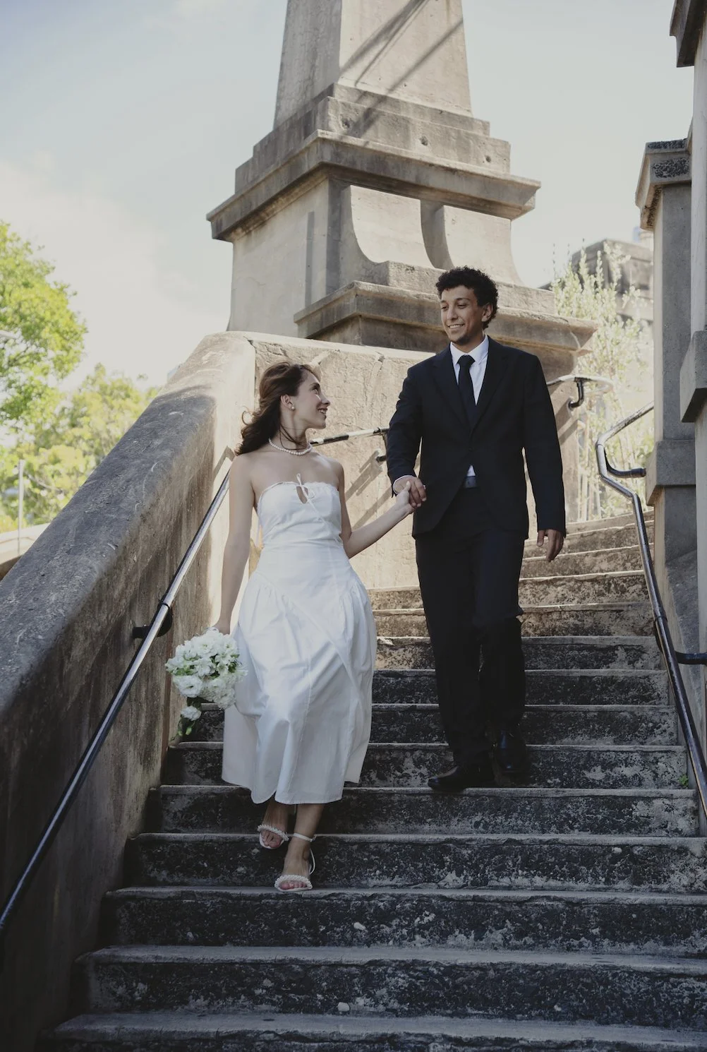 A bride and groom walking down outdoor stone stairs, holding hands and smiling at each other, with trees and a monument in the background.