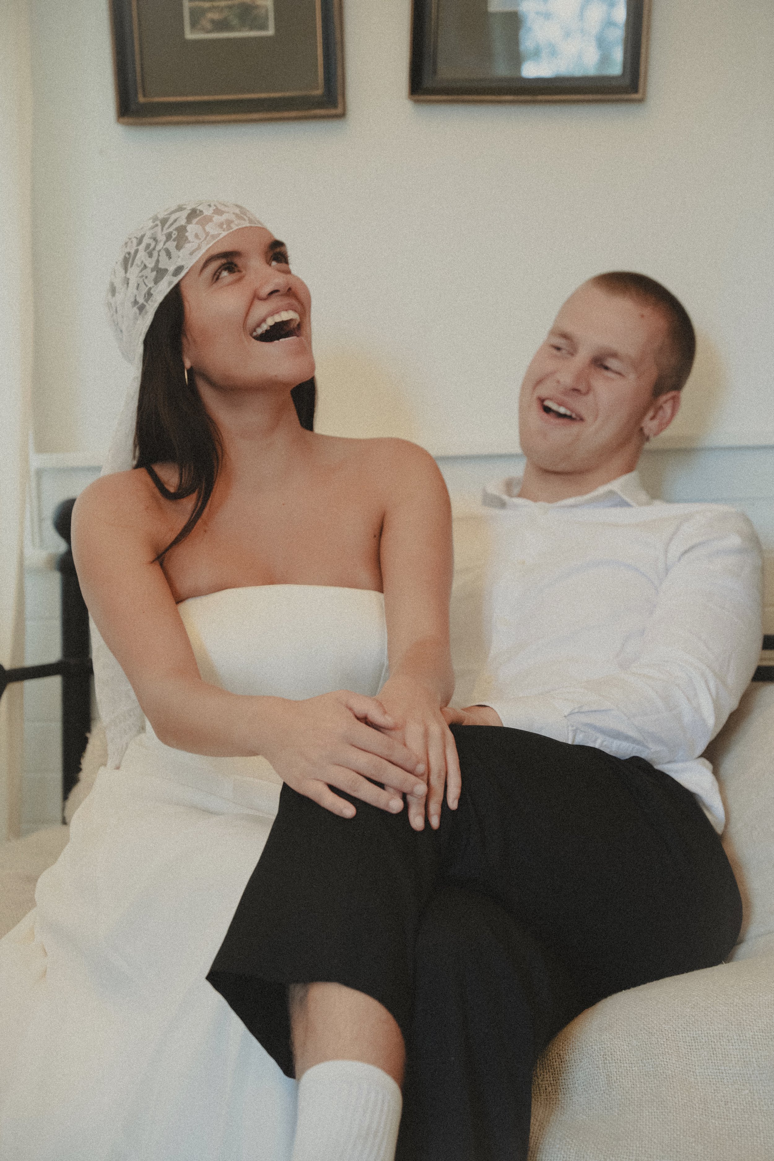 A woman wearing a lace headscarf and a strapless dress is sitting on a man's lap, both smiling and laughing, in a cozy room with framed pictures on the wall.