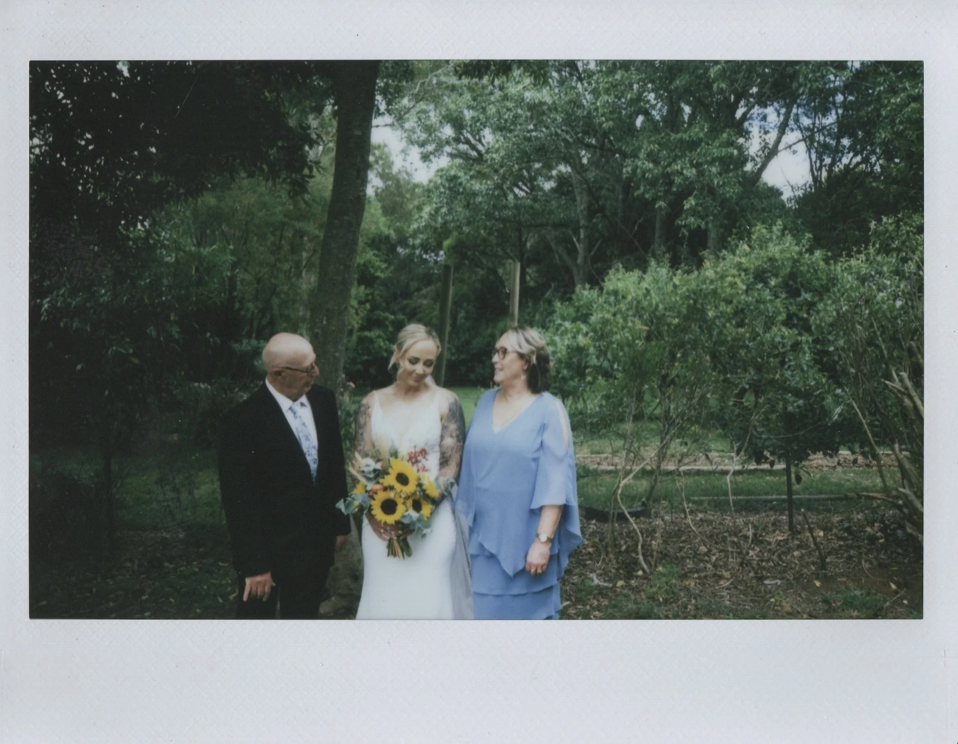 A woman in a white wedding dress holding a bouquet of sunflowers standing between an older man in a black suit and an older woman in a light blue dress, in a lush outdoor setting with trees and greenery.