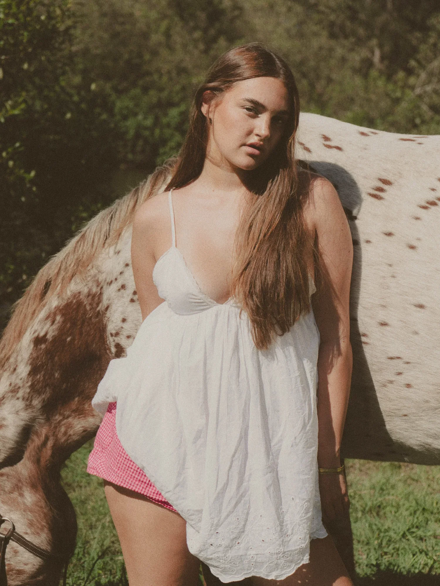 Young woman with long brown hair wearing a white dress standing outdoors next to a white horse with brown spots, green trees in the background.