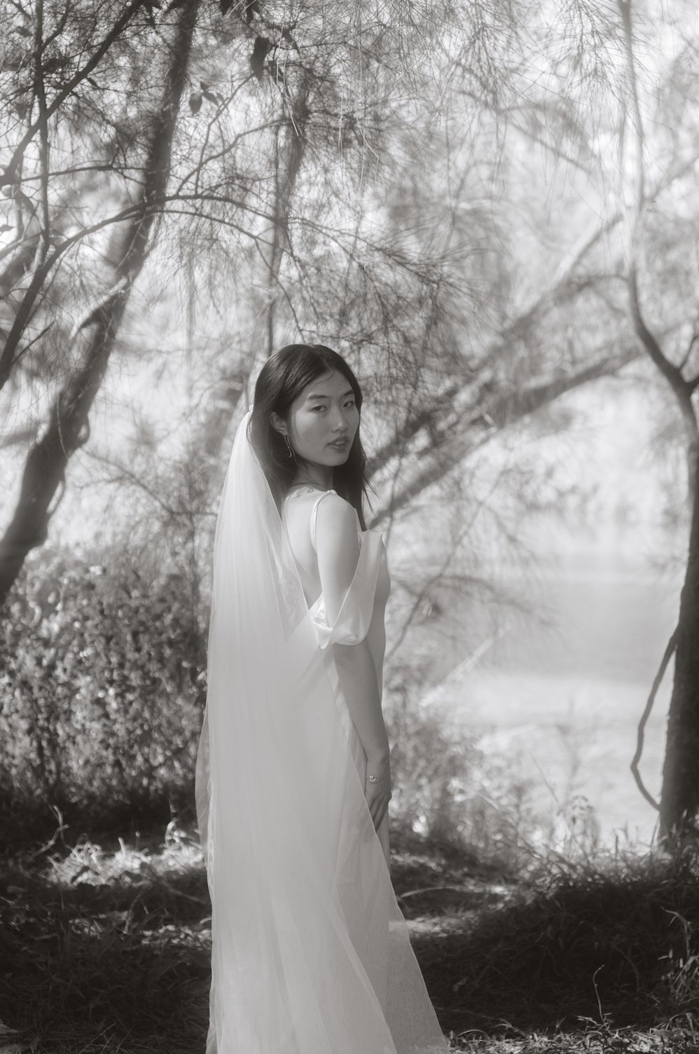 A woman standing outdoors among trees in a natural setting, wearing a flowing, light-colored dress and a veil, gazing over her shoulder.