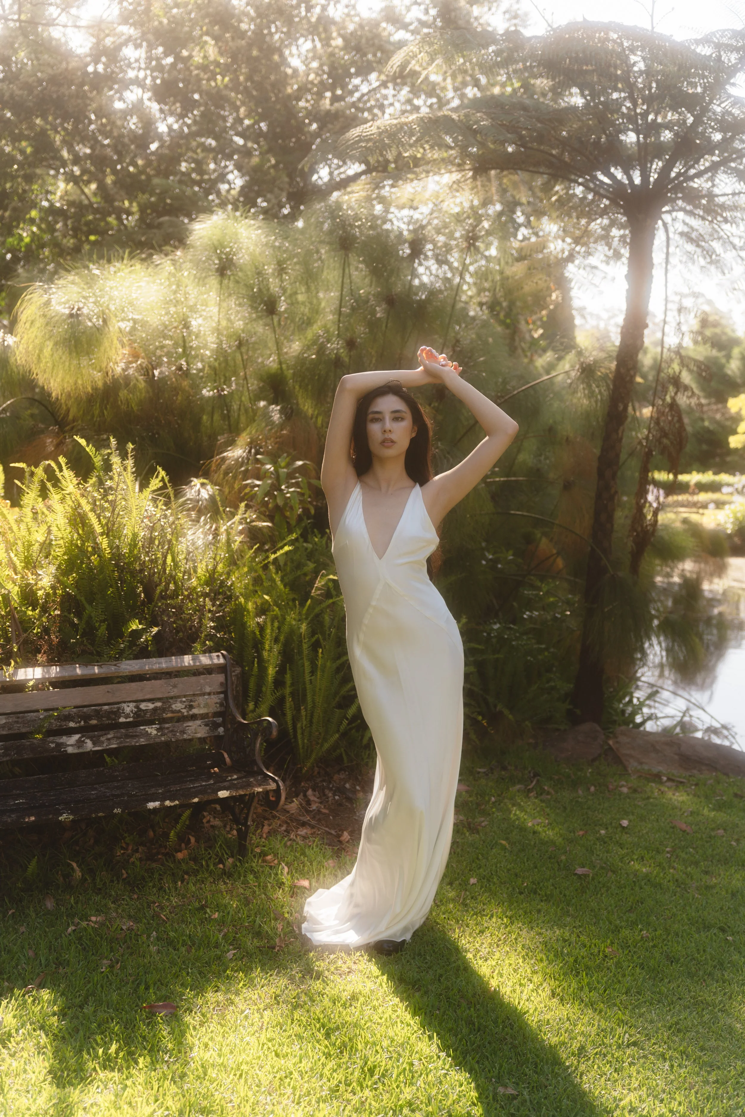 A woman in a white dress standing outdoors near a pond, with her arms raised above her head, surrounded by lush greenery and backlit by sunlight.